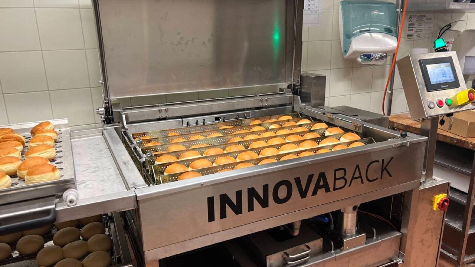 A commercial bakery machine labeled INNOVABACK is baking rows of evenly spaced bread rolls. A touchscreen control panel is visible on the right, and kitchen equipment is in the background.