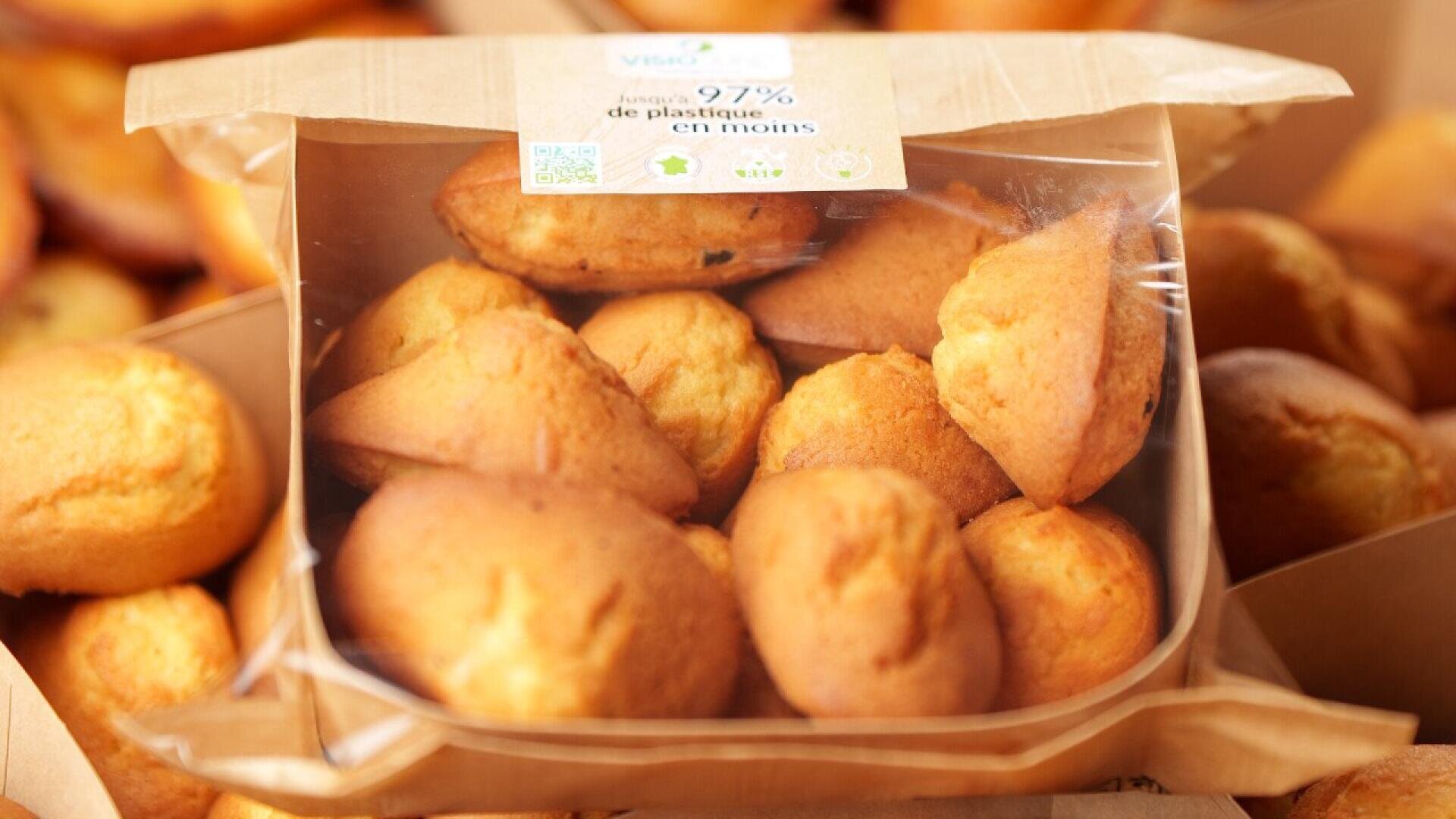 Eine durchsichtige Verpackung mit mehreren goldbraunen Madeleine-Kuchen ist ausgestellt, mit einem Etikett, das in französischer Sprache auf den reduzierten Plastikverbrauch hinweist. Im Hintergrund sind weitere Madeleines zu sehen.