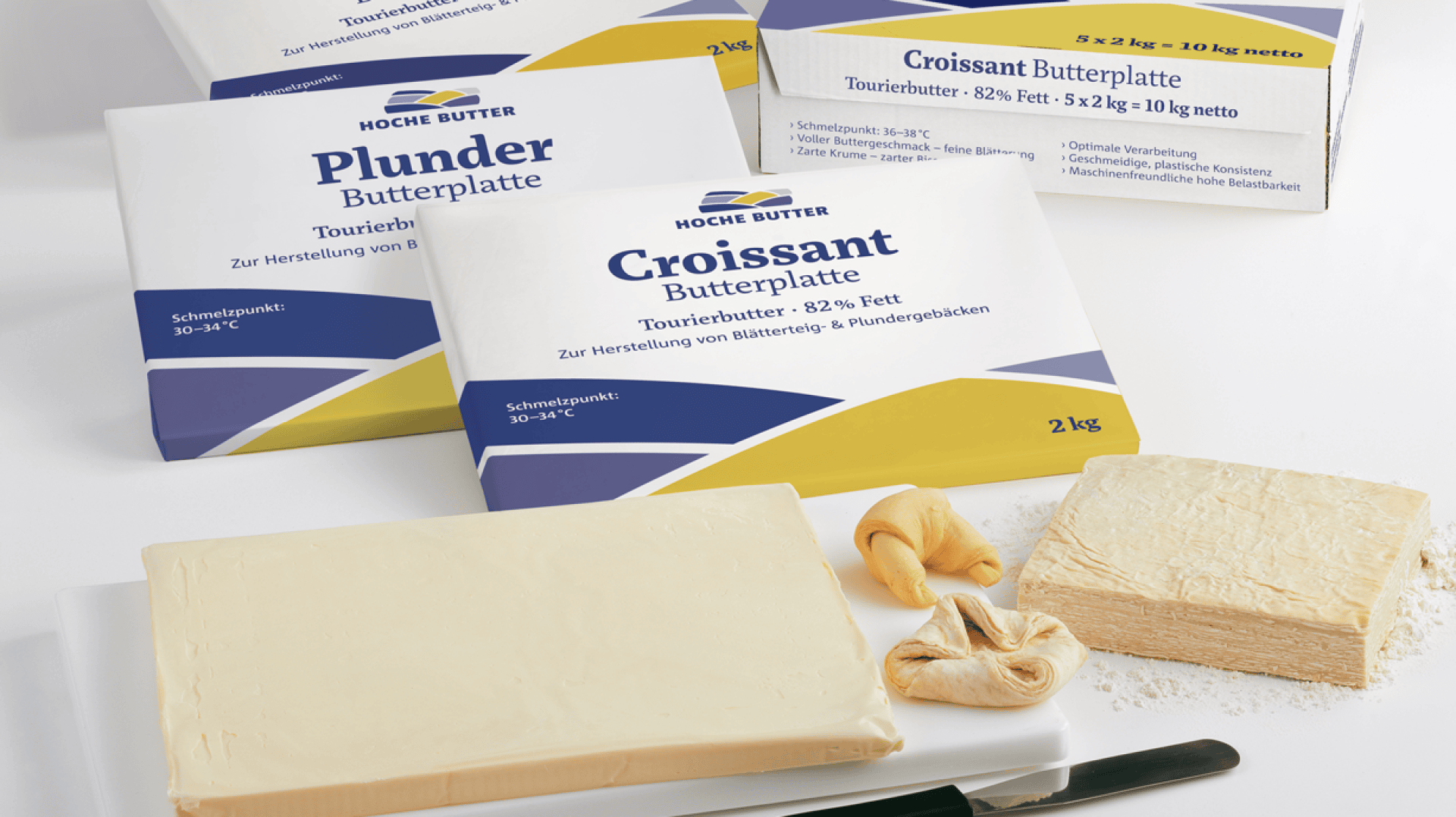 Butterblöcke für Croissants und Plundergebäck sind mit ausgepackter Butter, Blätterteig und geformtem Teig auf einem weißen Schneidebrett abgebildet, daneben liegt ein Messer.