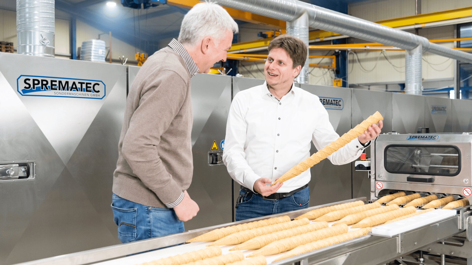 Zwei Männer in einer Fabrik inspizieren lange Gebäckrollen auf einem Fließband. Beide lächeln und einer hält ein Gebäckstück in der Hand. Im Hintergrund sind industrielle Backmaschinen mit der Aufschrift SPREMATEC zu sehen.