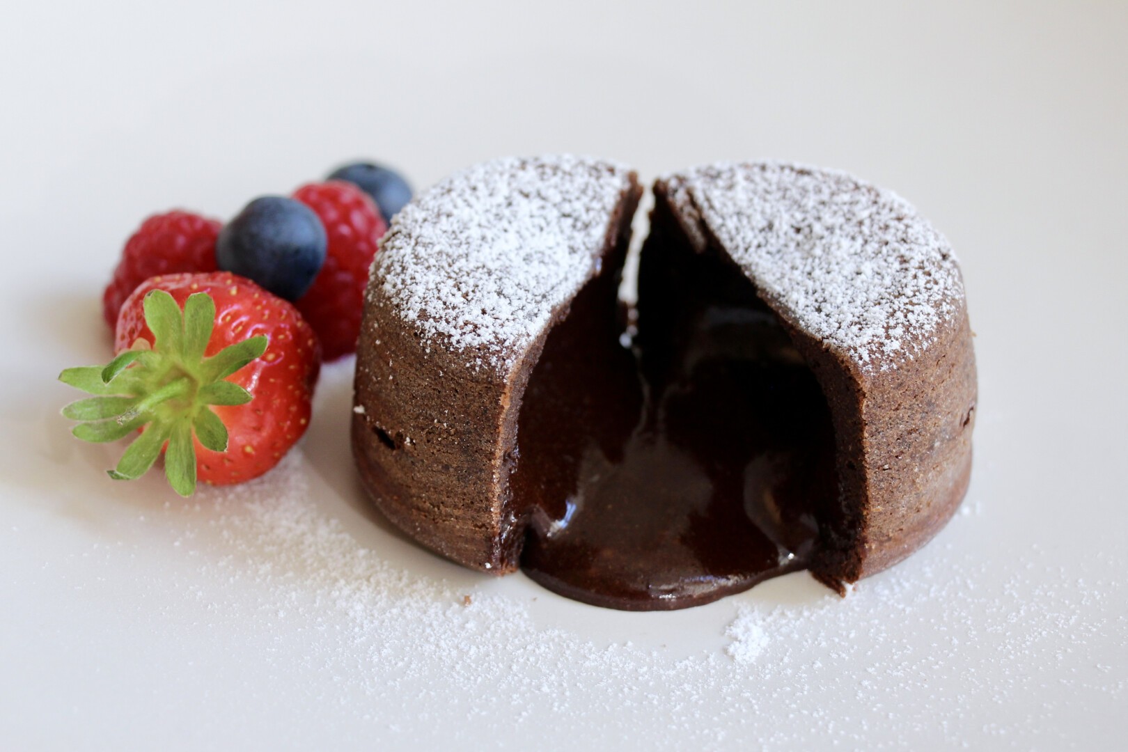 A chocolate lava cake dusted with powdered sugar is cut open, with molten chocolate oozing out. Fresh strawberries, raspberries, and blueberries are placed beside the dessert on a white plate.