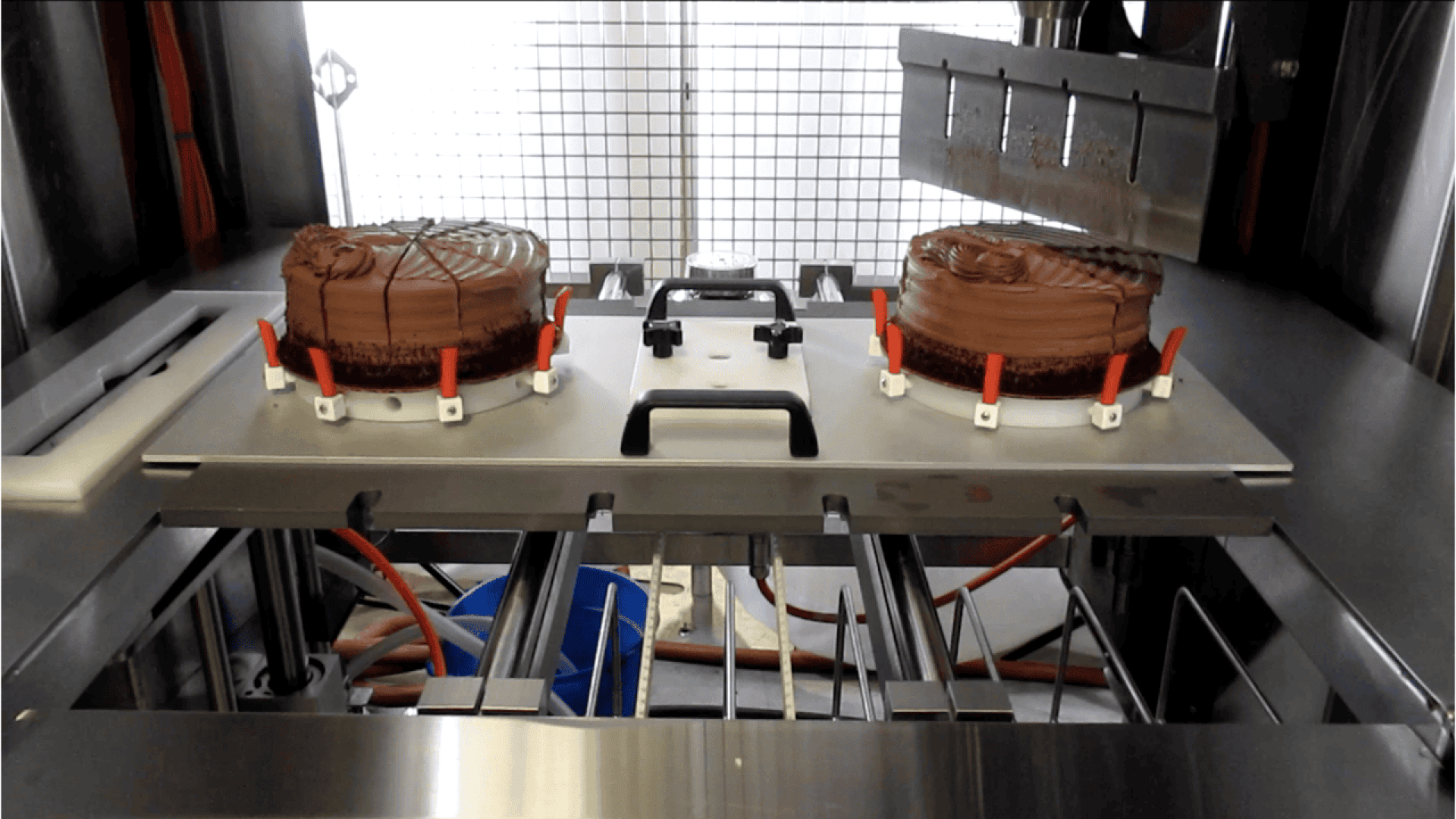 Zwei Schokoladenkuchen, die auf einem Metalltablett mit Klammern platziert sind, befinden sich unter einer Schneidemaschine in einer industriellen Lebensmittelverarbeitungsanlage.