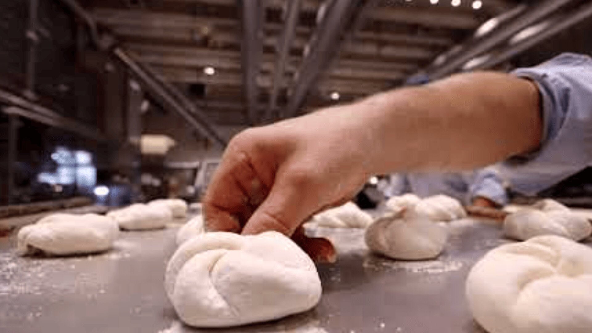 A close-up of a hand shaping pieces of dough into knots on a floured surface in an industrial kitchen or bakery setting.