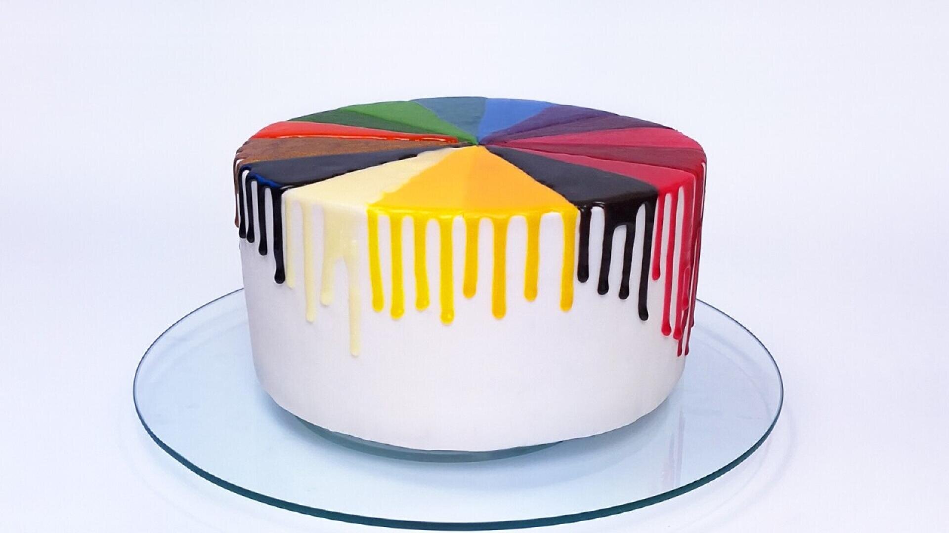 Ein runder Kuchen mit weißen Seiten und bunten dreieckigen Abschnitten auf der Oberseite, jeder mit farbigem Zuckerguss, der an den Seiten heruntertropft, ausgestellt auf einer Glasplatte vor einem weißen Hintergrund.