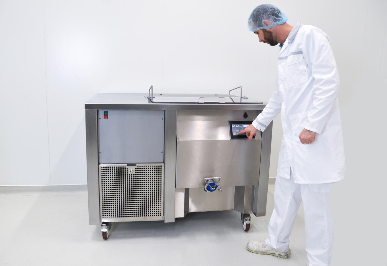 A man in a white lab coat, hairnet, and shoe covers operates a stainless steel industrial machine with a digital control panel in a clean, white room.