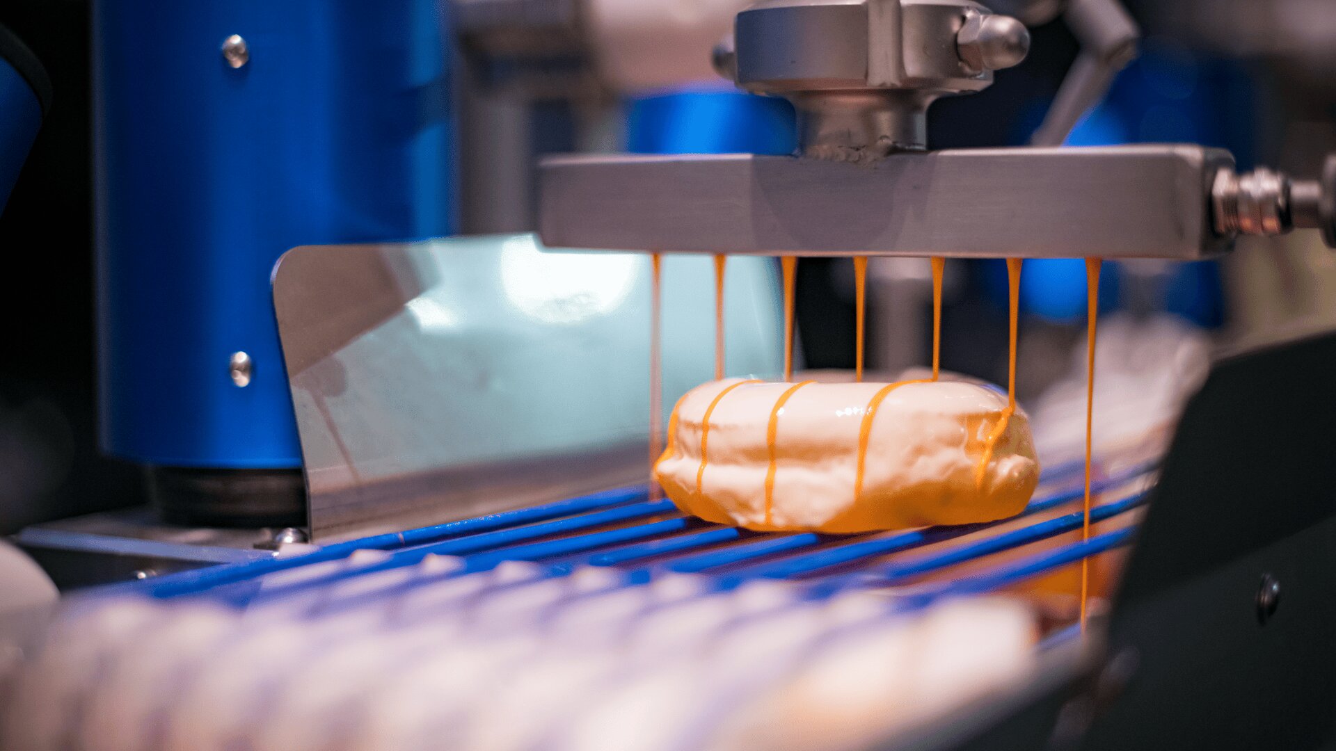 Ein Donut am Fließband wird von einer Maschine in einer Fabrik mit Zuckerguss glasiert, der gleichmäßig auf das Gebäck tropft.