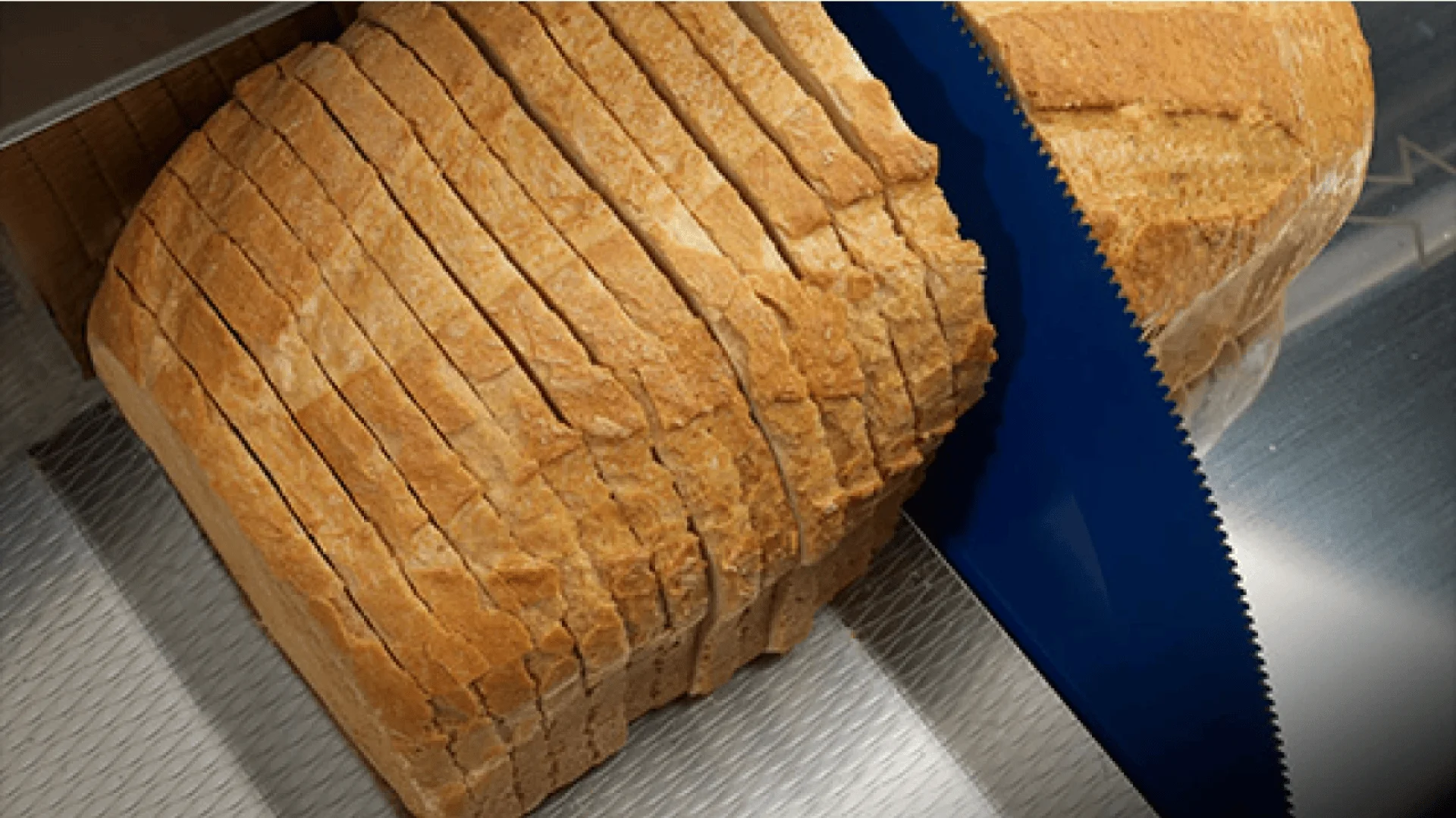 Nahaufnahme eines Brotlaibs, der mit einer großen gezahnten Brotschneidemaschine geschnitten wird, wobei gleichmäßige Scheiben und Krümel auf der Schnittfläche zu sehen sind.