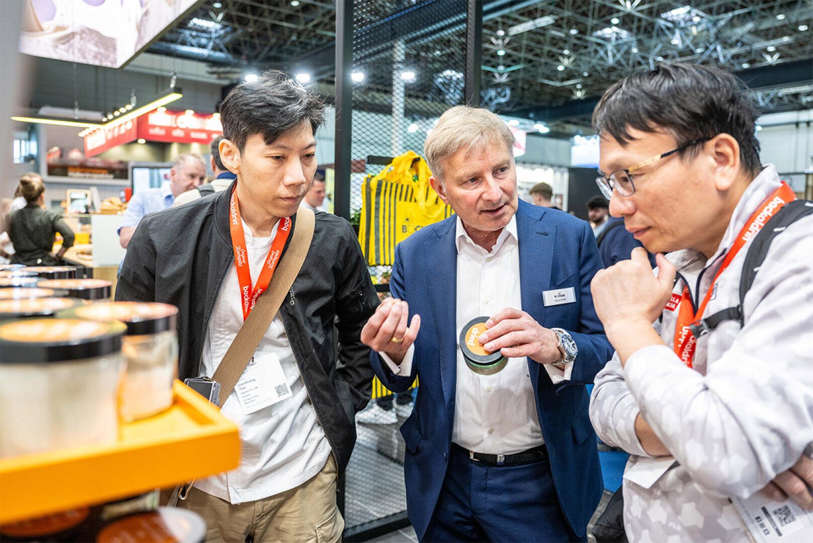 Three men stand together at a trade show or exhibition booth, discussing a product. One man in a blue suit is holding a small container and gesturing, while the other two listen intently with red lanyards around their necks.