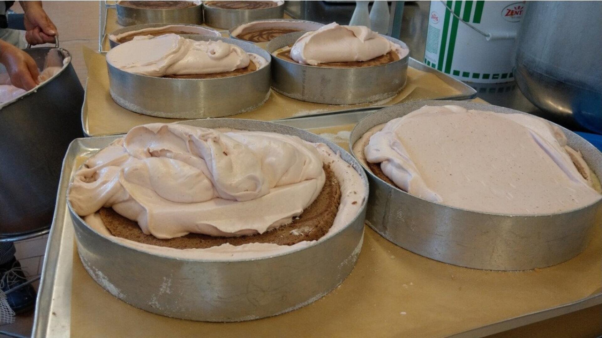 Several round cakes in metal pans are topped with generous dollops of light pink frosting, awaiting further decoration or spreading. The cakes are on a counter in a commercial kitchen setting.
