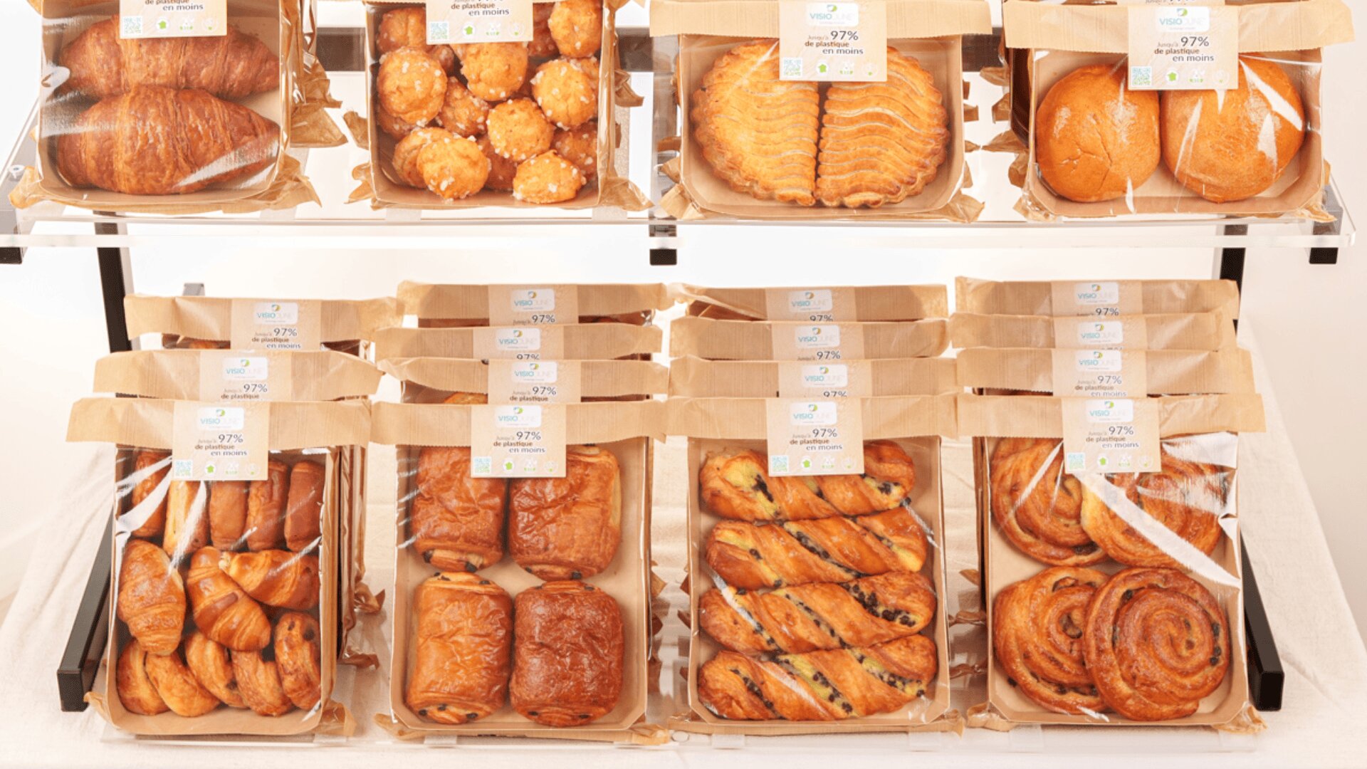 Assorted pastries and breads arranged in paper trays on display shelves, including croissants, chocolate breads, swirled buns, and other baked goods, each labeled with price tags.