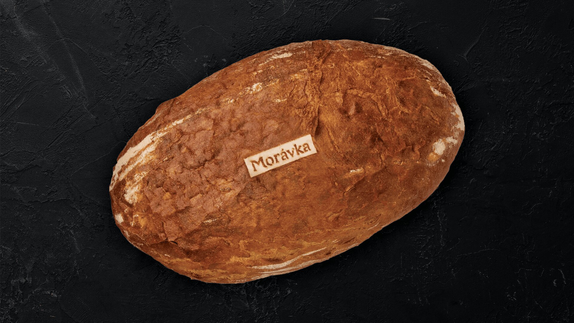 Ein runder Laib Schwarzbrot mit einem kleinen weißen Etikett mit der Aufschrift Morávka auf der Oberseite, auf einem dunkel strukturierten Hintergrund.