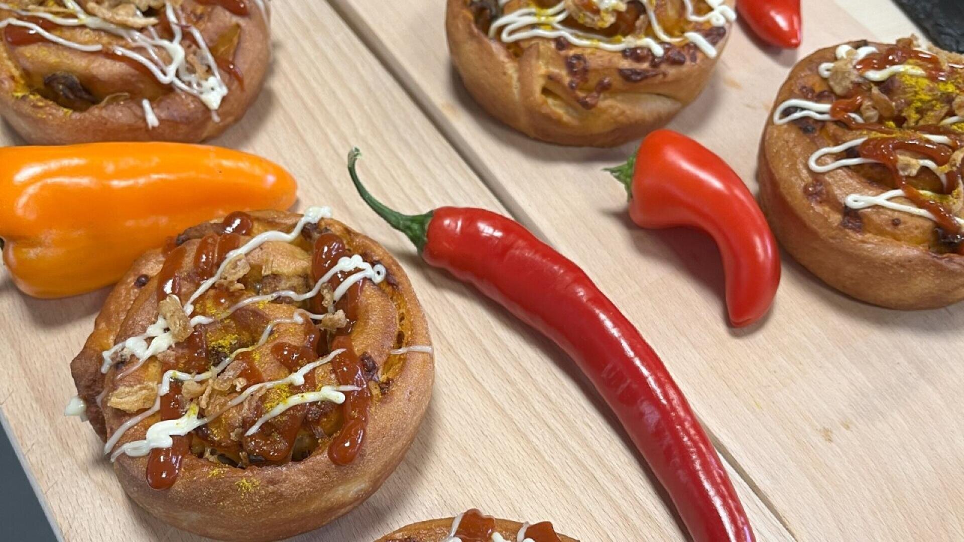 Spicy bread rolls topped with sauces and onions, displayed on a wooden board alongside whole orange and red chili peppers.