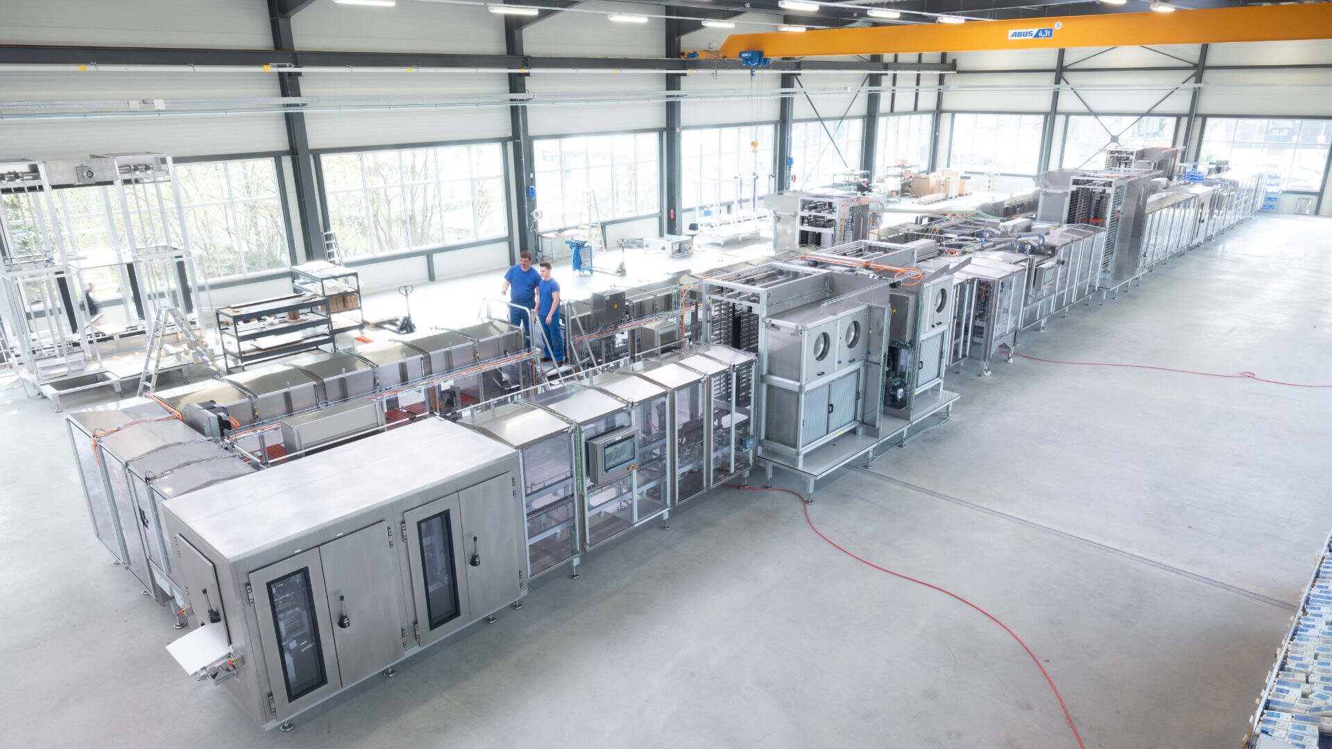 Eine große Industrieanlage mit zwei Personen, die ein langes, komplexes Fließband mit Maschinen aus rostfreiem Stahl inspizieren, beleuchtet von natürlichem Licht aus hohen Fenstern.