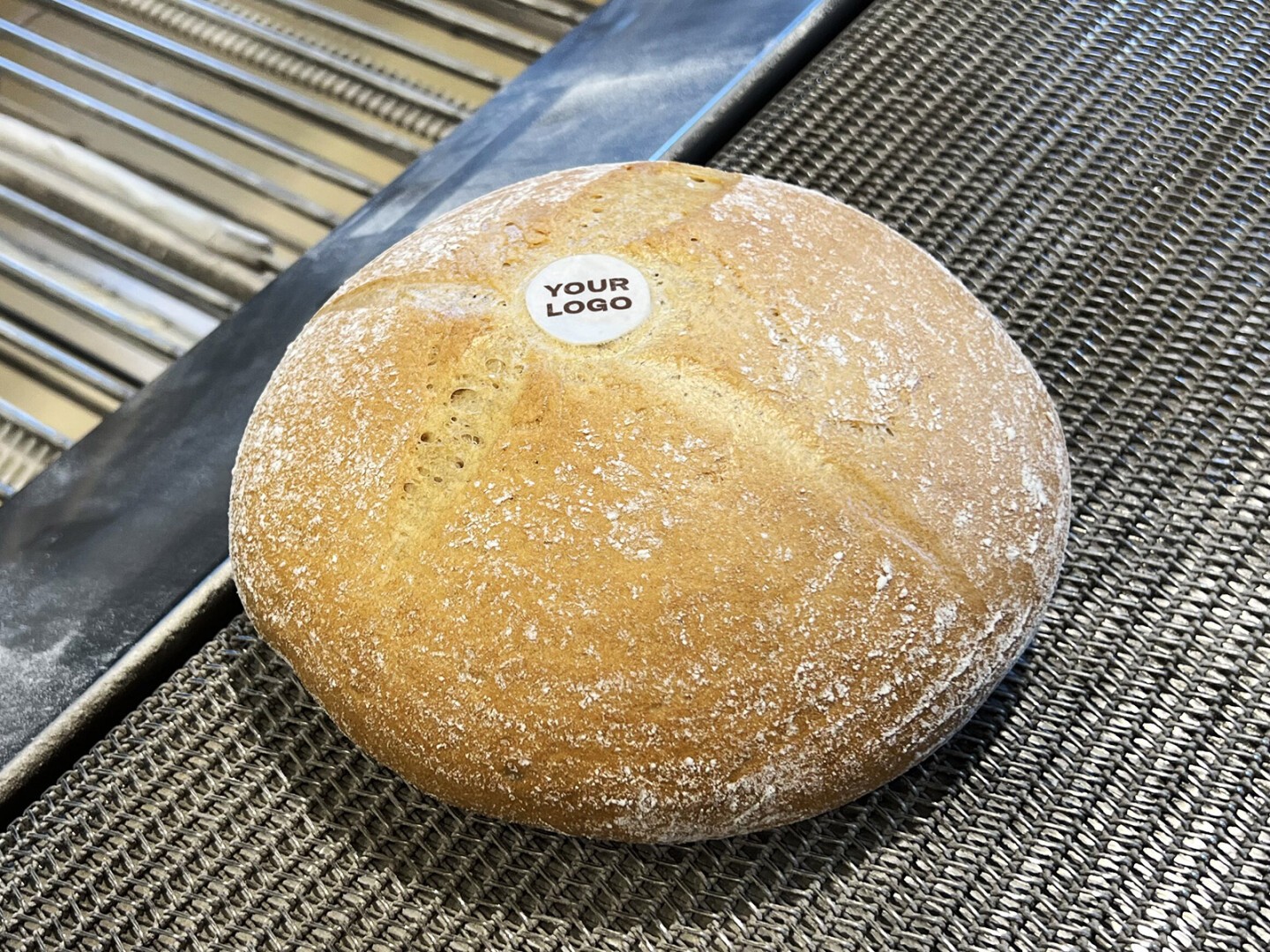A round loaf of bread sits on a conveyor belt, topped with a small white sticker that reads YOUR LOGO.