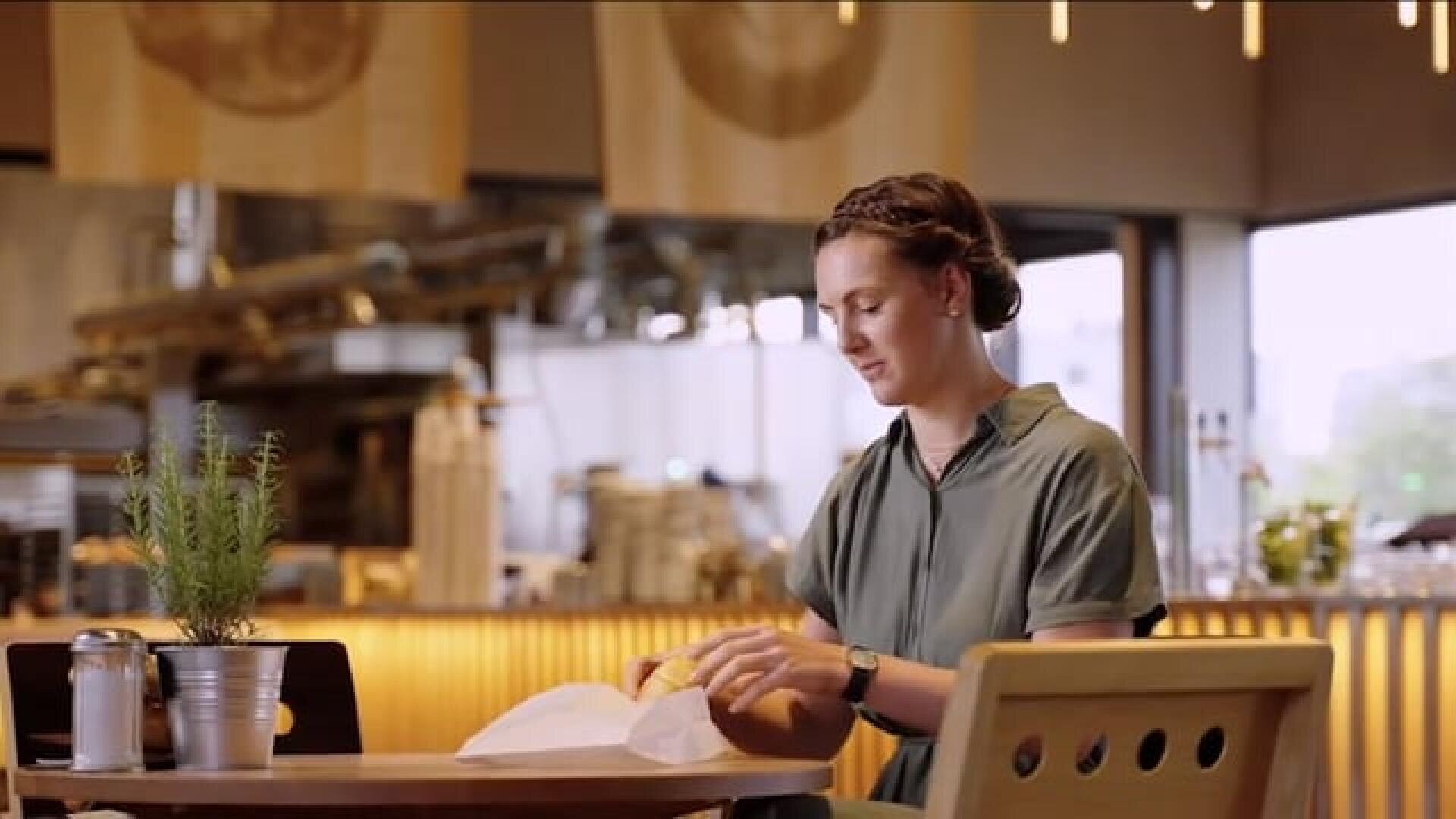 A woman with braided hair sits alone at a table in a modern café, unwrapping food. There is a potted plant, salt shaker, and napkin holder on the table. The background shows a bright, open kitchen area.