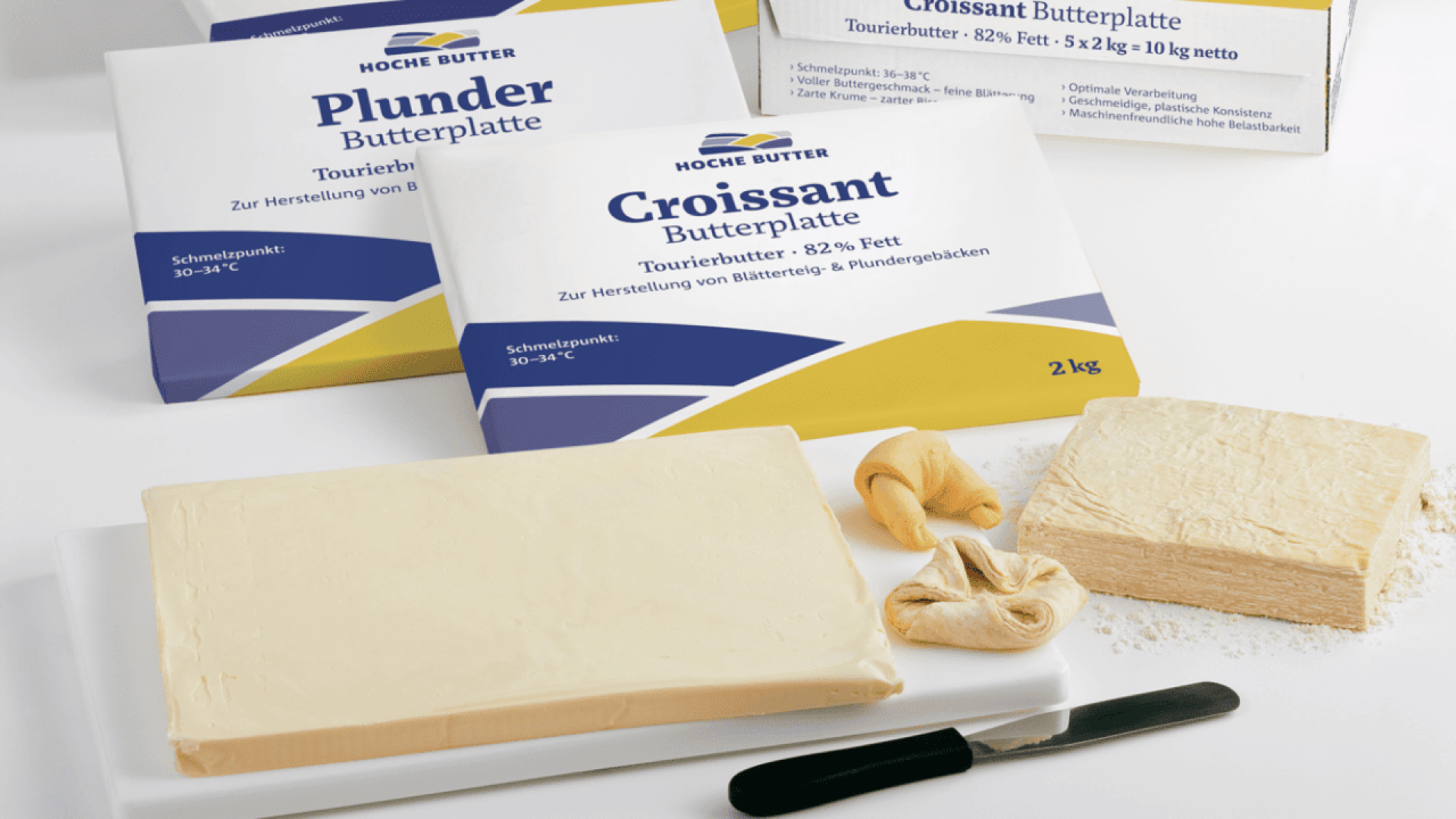 Rectangular blocks of butter labeled Plunder Butterplatte and Croissant Butterplatte are placed near raw dough, small unbaked croissants, and a butter knife on a white surface.