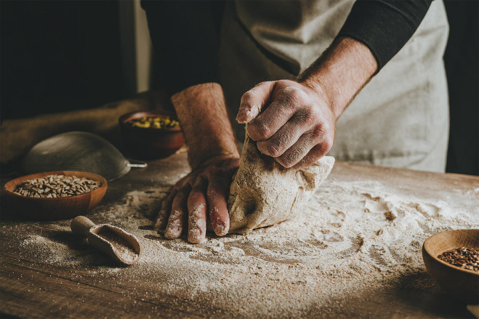 A person kneads dough on a floured wooden surface, surrounded by small bowls of seeds and grains. Their hands and arms are dusted with flour, and they are wearing a light apron.
