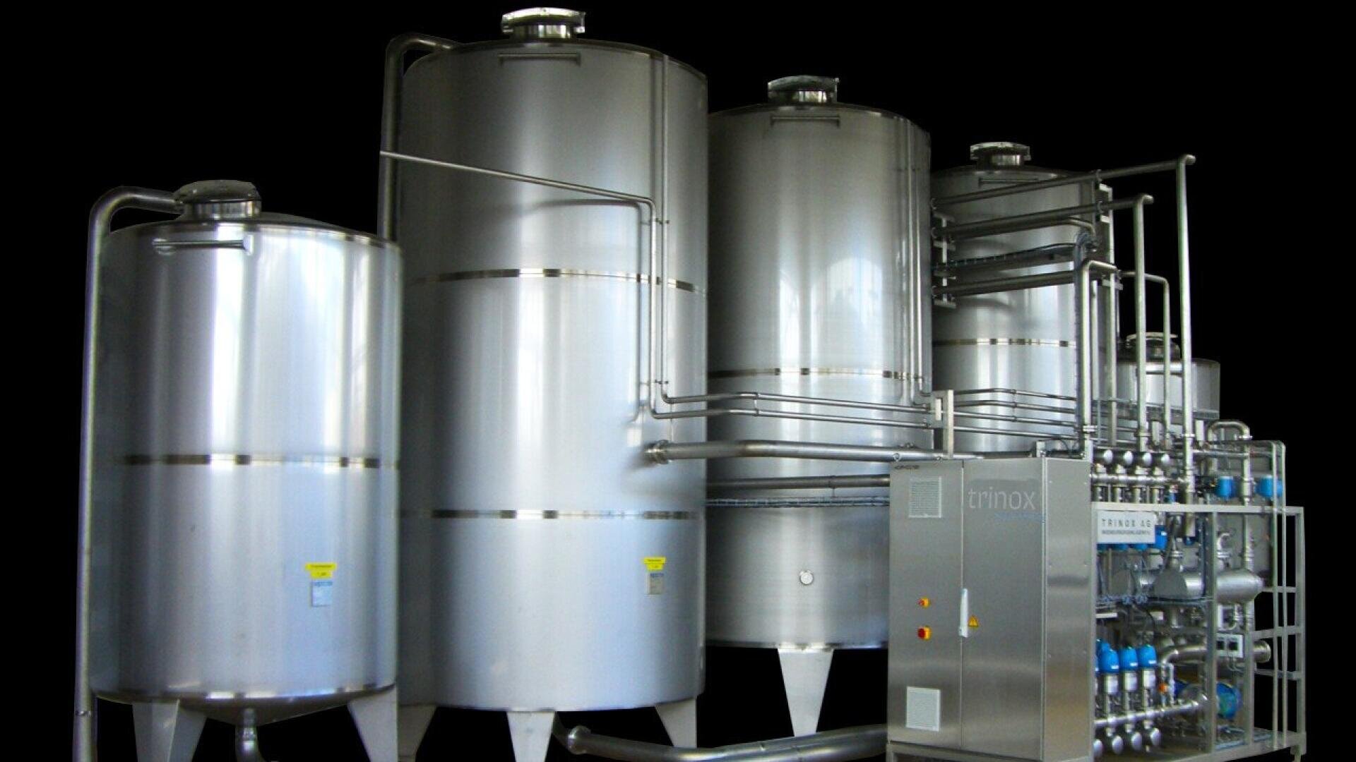 A row of large, shiny stainless steel industrial tanks connected by pipes and machinery, used for processing or storing liquids, set against a black background.