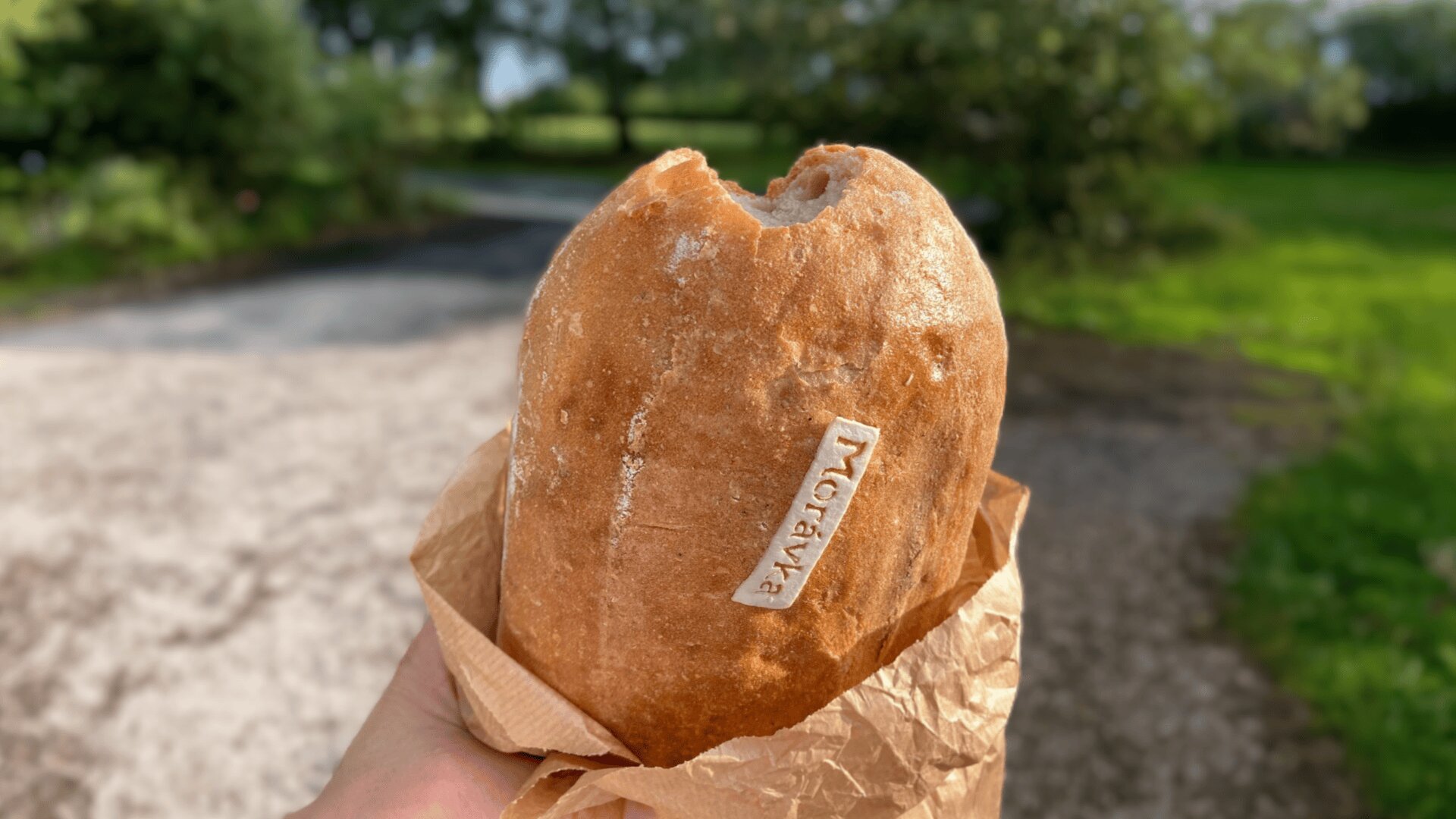 Eine Hand hält einen teilweise gegessenen Laib Brot, der in braunes Papier eingewickelt ist und ein kleines Etikett auf der Kruste trägt. Der Hintergrund zeigt eine verschwommene Szene im Freien mit Grünpflanzen und einem Kiesweg.