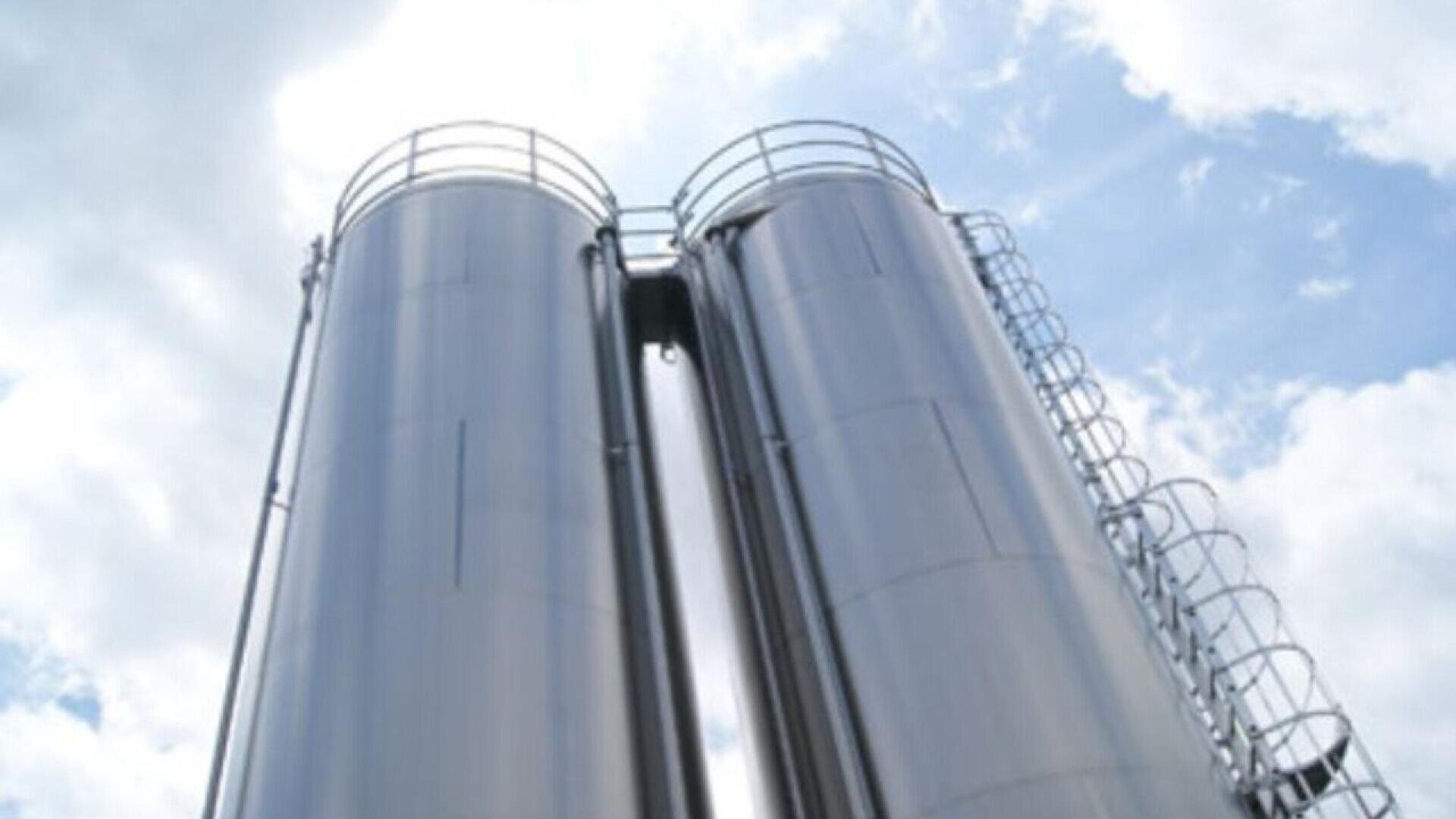 Two tall, shiny metal silos stand side by side against a partly cloudy sky, each with a circular platform and ladder running along the side.