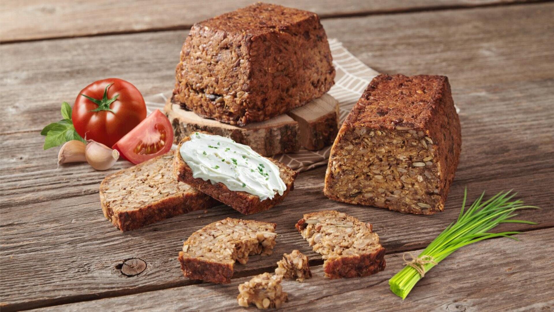 Aufgeschnittenes Mehrkornbrot auf einem Holztisch, eine Scheibe ist mit Frischkäse und Schnittlauch bestreut. Um das Brot herum liegen Tomatenhälften, Knoblauch und frischer Schnittlauch, was einen rustikalen, natürlichen Rahmen schafft.