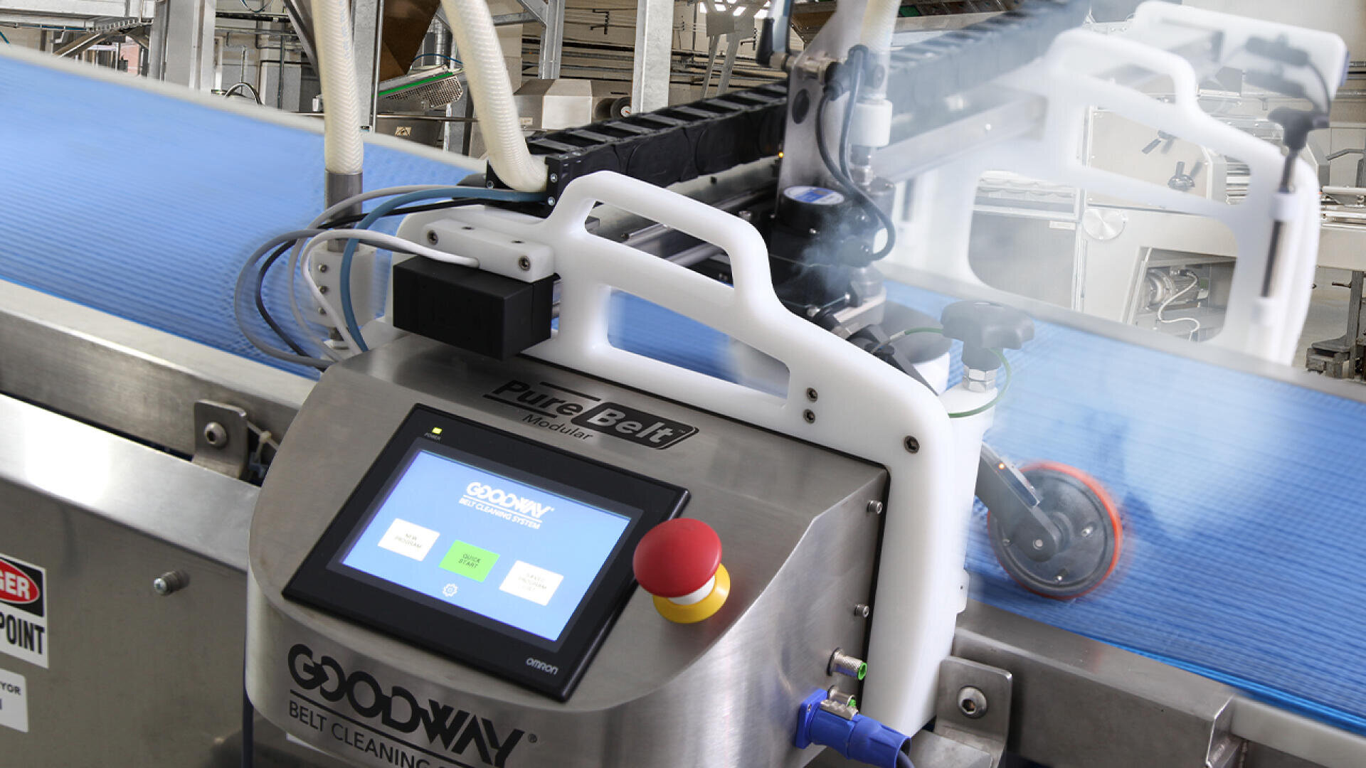 A conveyor belt cleaning machine with a touchscreen display and emergency stop button operates on a blue conveyor belt in an industrial setting. Nearby machinery and pipes are visible in the background.