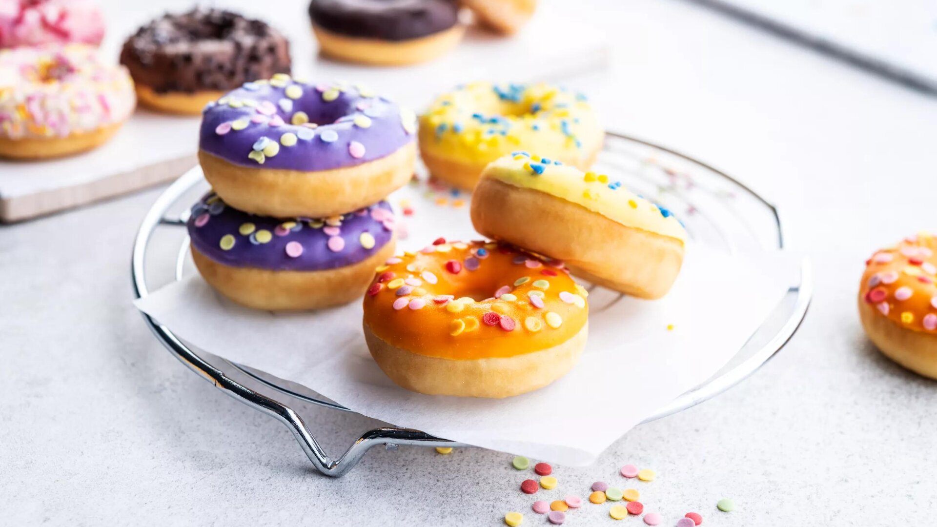 Bunte Donuts mit leuchtendem lila, orangefarbenem und gelbem Zuckerguss und Streuseln sind auf einem mit Pergamentpapier ausgelegten Drahtgestell gestapelt, während im Hintergrund weitere verzierte Donuts zu sehen sind.