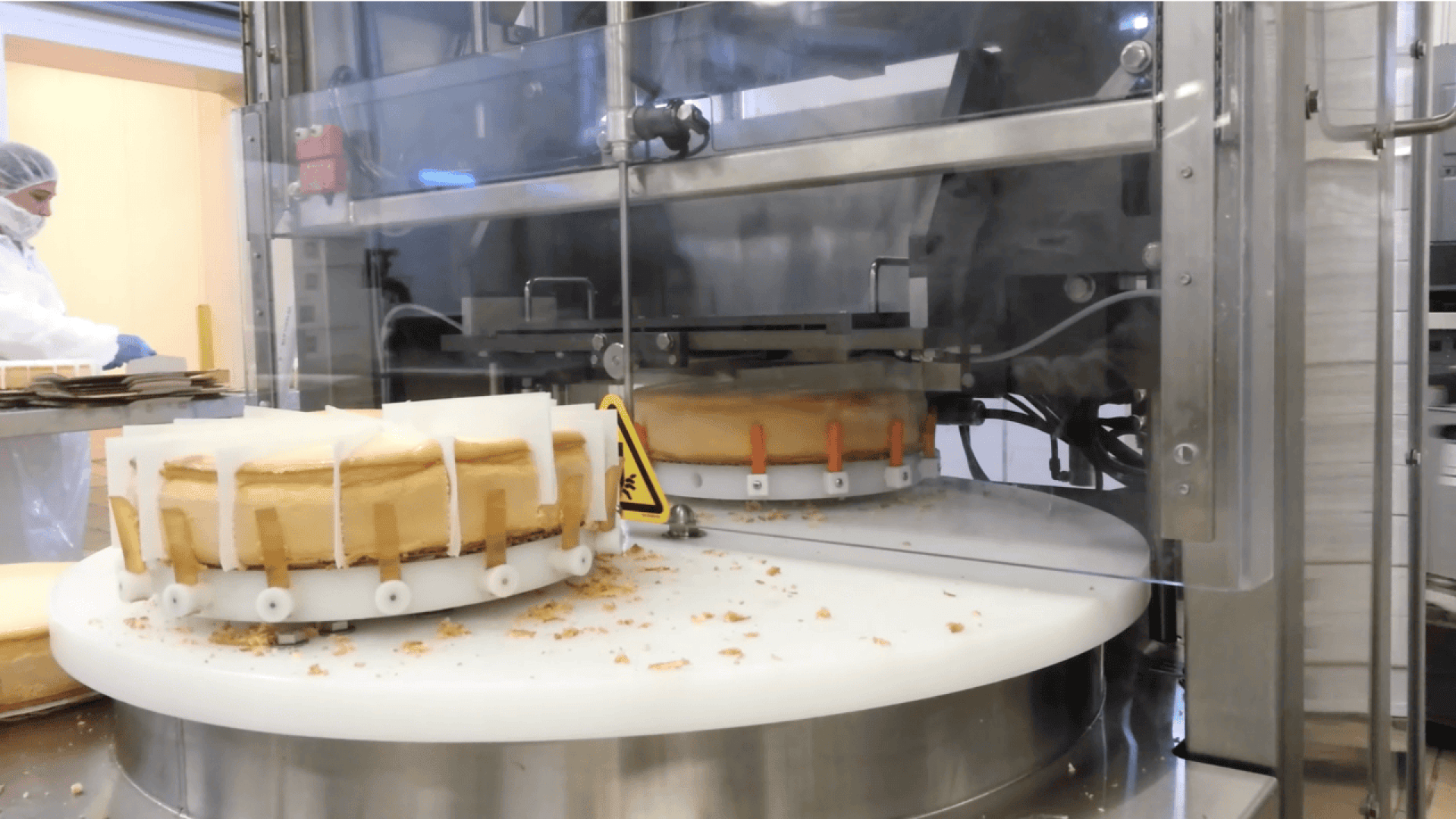 Nahaufnahme einer Käsekuchenschneidemaschine in einer Fabrik, die Kuchen in gleichmäßige Portionen schneidet, während ein Arbeiter in Schutzkleidung im Hintergrund zusieht.
