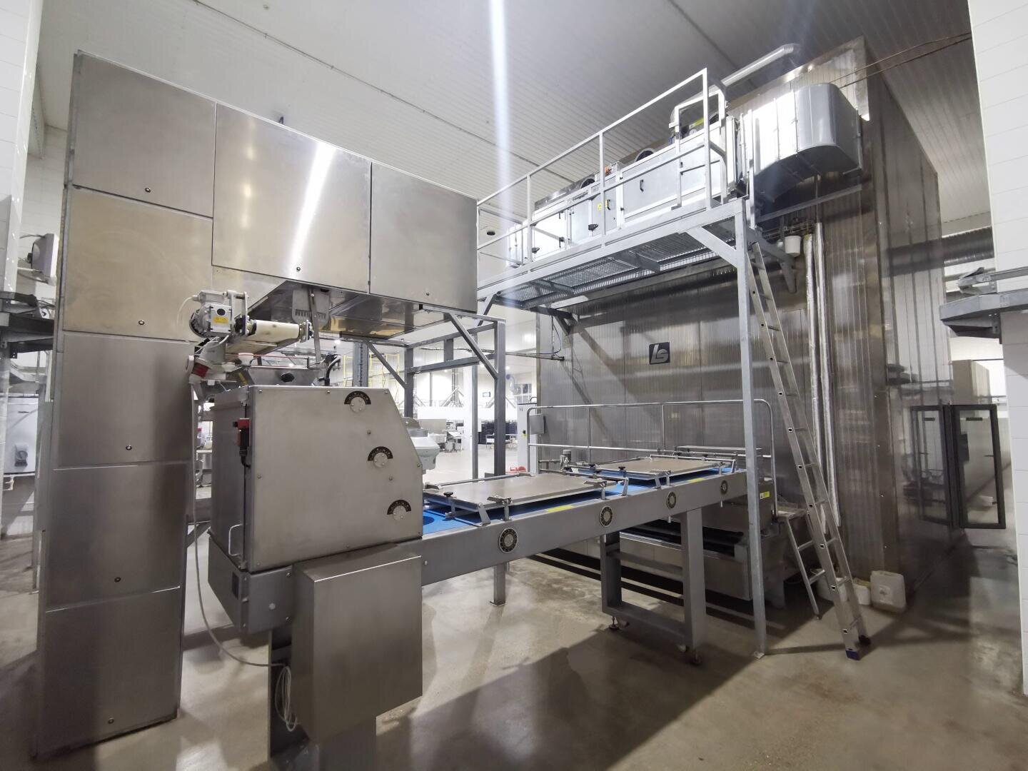 Industrial food processing equipment in a clean, modern factory setting, featuring stainless steel machinery, conveyor belts, a raised platform with railings, and a metal ladder. The floor is shiny and the area is well-lit.
