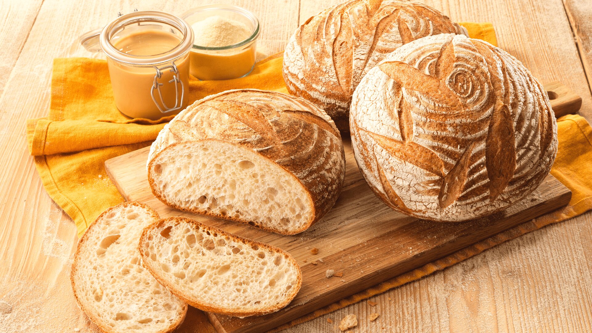 Auf einem Holzbrett liegen runde Brote aus handwerklich hergestelltem Sauerteig mit dekorativer Kruste, zwei Scheiben sind abgeschnitten und liegen davor. Gläser mit Aufstrich und ein gelbes Tuch stehen in der Nähe auf einem rustikalen Holztisch.