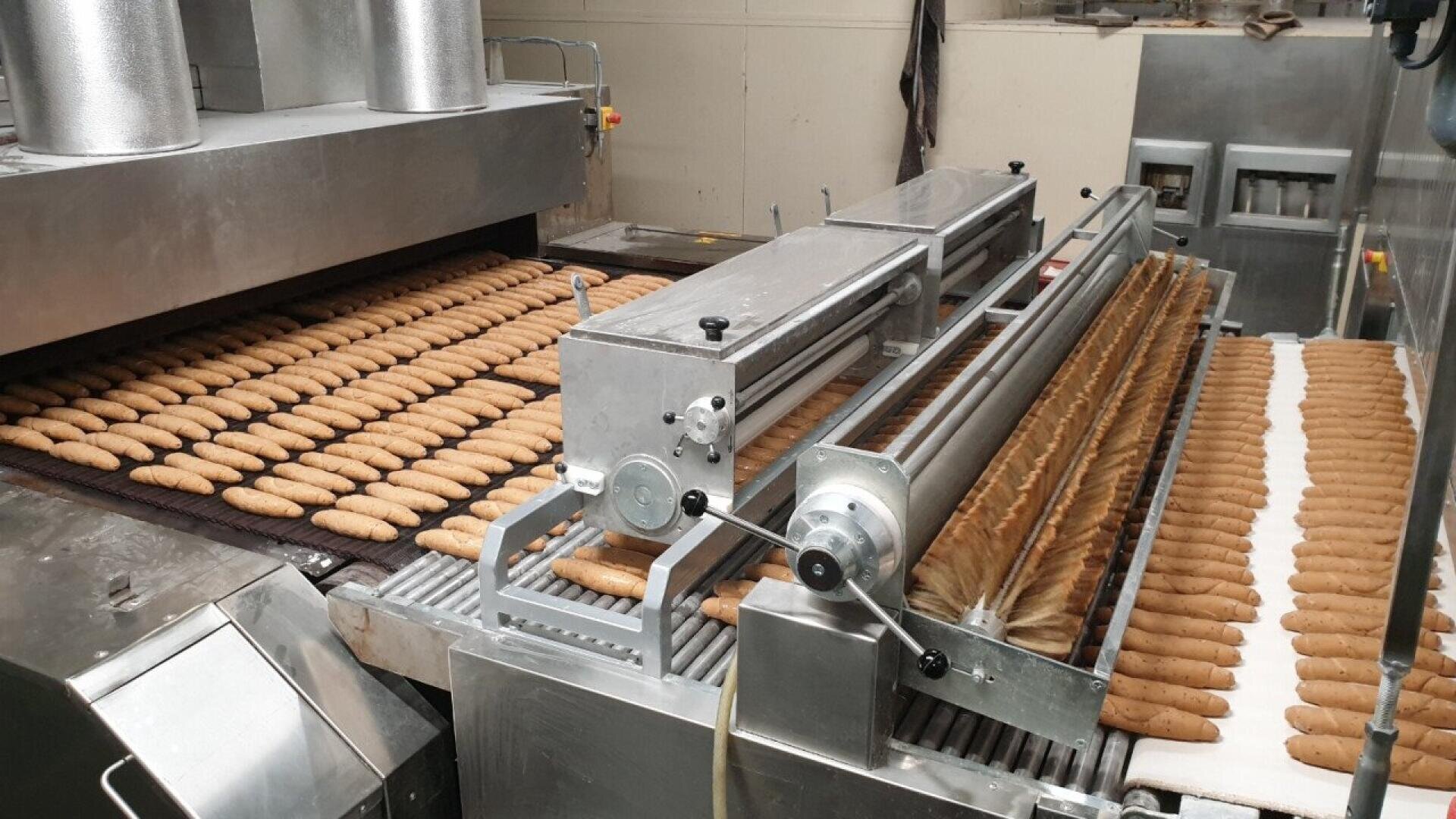 In einer Großbäckerei laufen Reihen von Brotstangen oder Baguettes über ein Förderband, das durch Metallmaschinen und Öfen zur automatischen Verarbeitung und zum Backen führt.