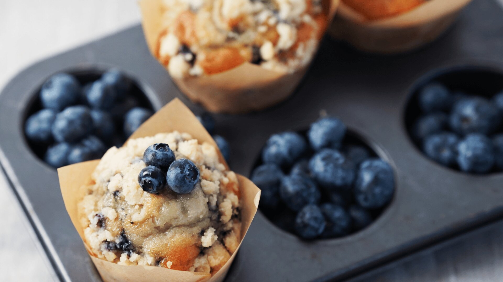 Eine Nahaufnahme von Blaubeermuffins in einer Backform, mit frischen Blaubeeren in leeren Muffinförmchen. Die Muffins sind mit einer Streuselschicht und zusätzlichen Blaubeeren belegt.