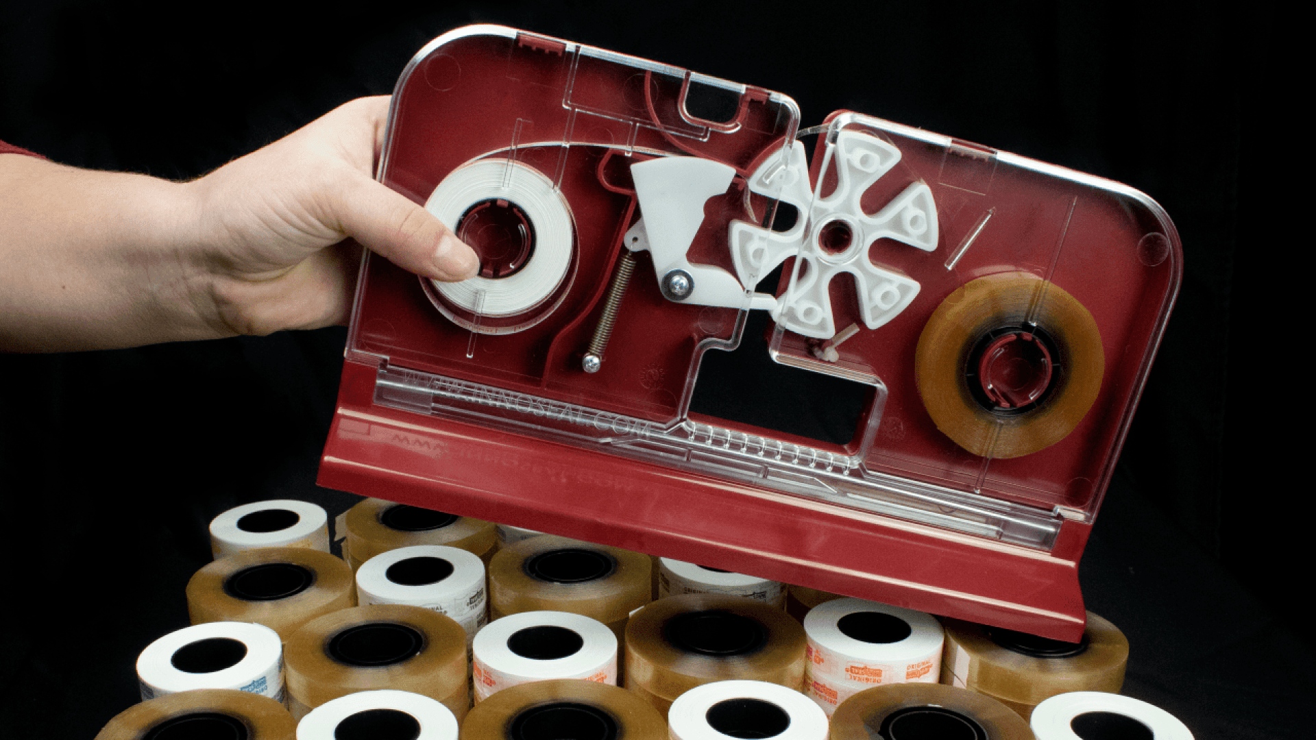 Eine Hand hält einen roten Klebebandabroller mit geöffnetem Deckel, der den inneren Mechanismus zeigt. Darunter sind mehrere Rollen durchsichtiges und weißes Klebeband vor einem schwarzen Hintergrund gestapelt.