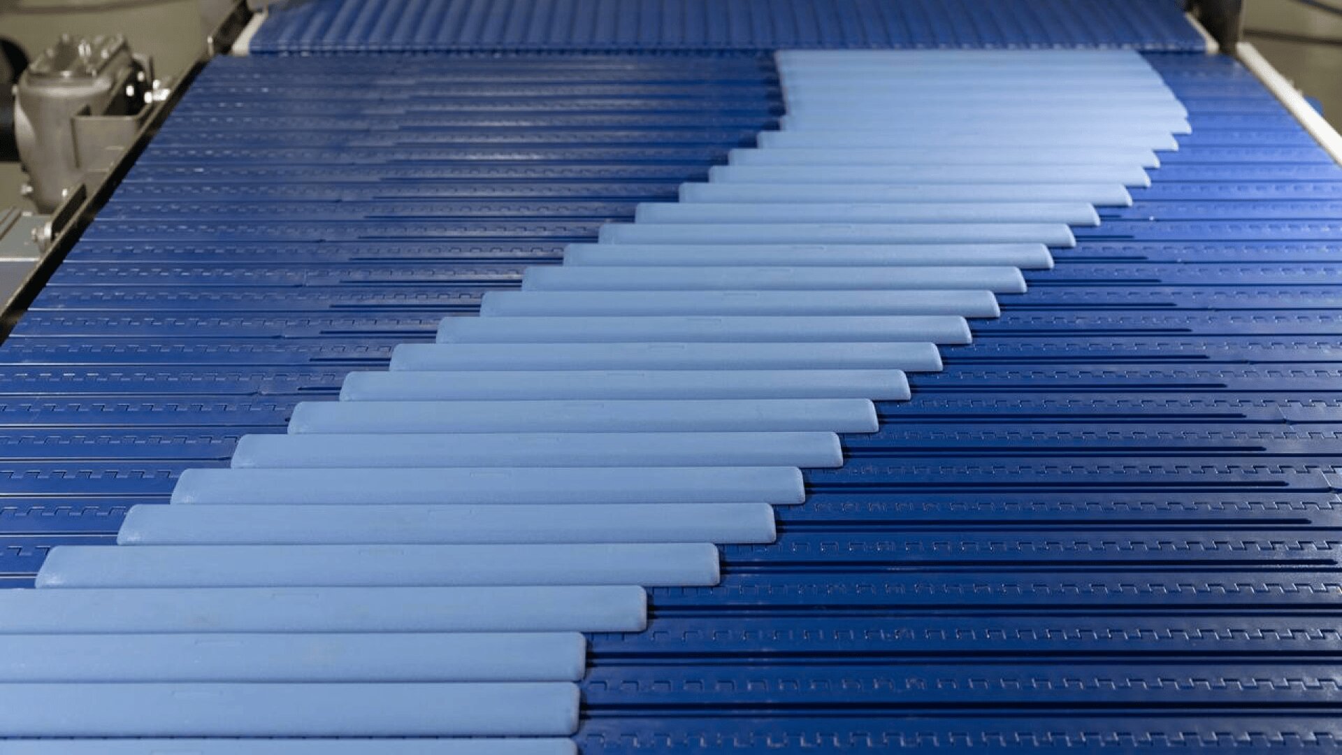 A conveyor belt with overlapping blue rectangular items moving diagonally across a textured blue surface in a factory or industrial setting.