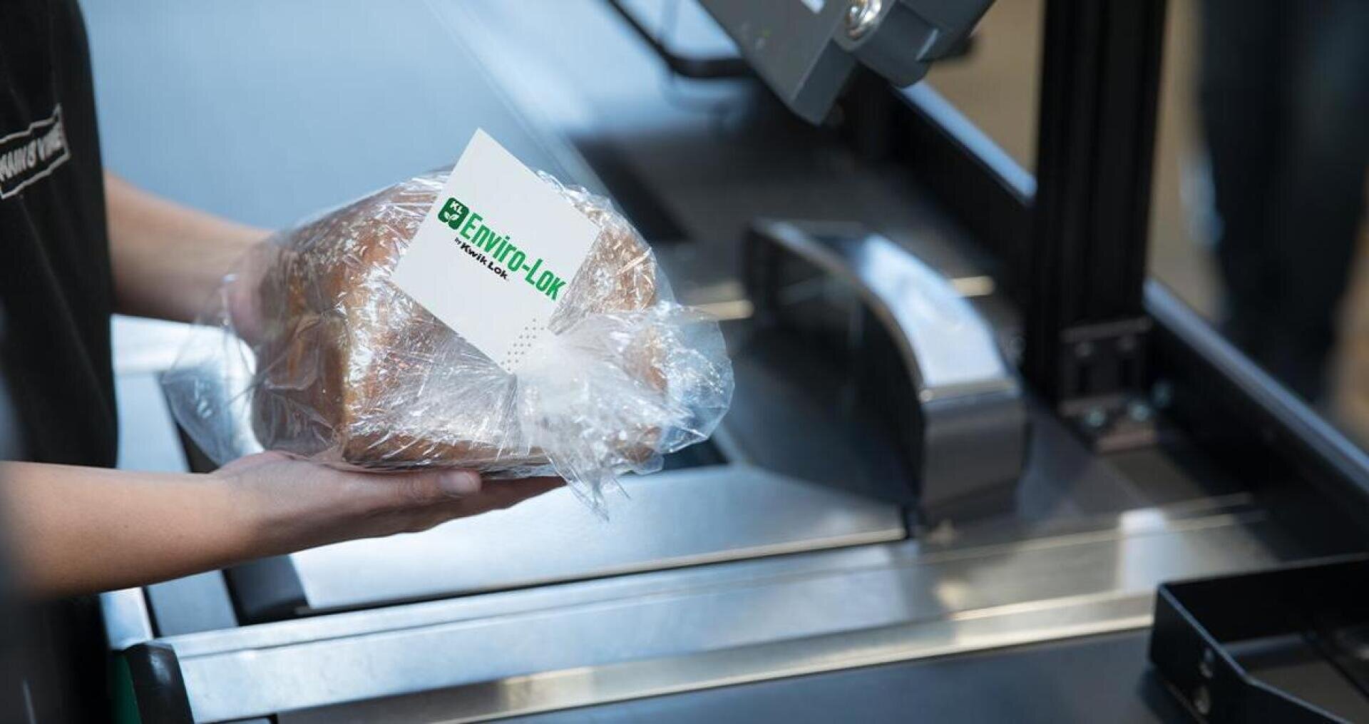 Eine Person hält ein in Plastik eingewickeltes Brot mit einem Envirolok-Etikett und bereitet sich darauf vor, es an einer Selbstbedienungskasse zu scannen.