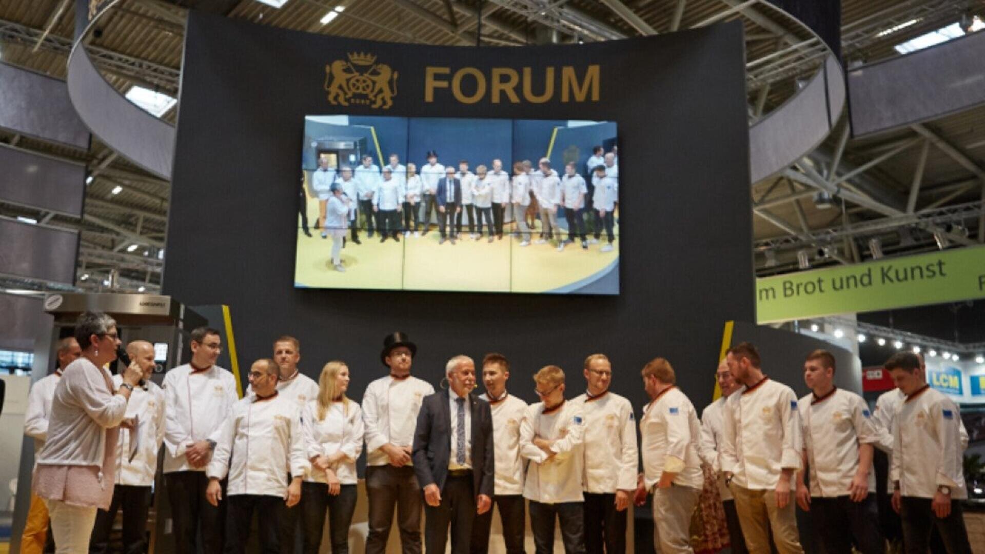 A group of chefs and presenters stand on a stage under a large screen displaying a photo of themselves; the backdrop reads FORUM in large yellow letters at an indoor event.
