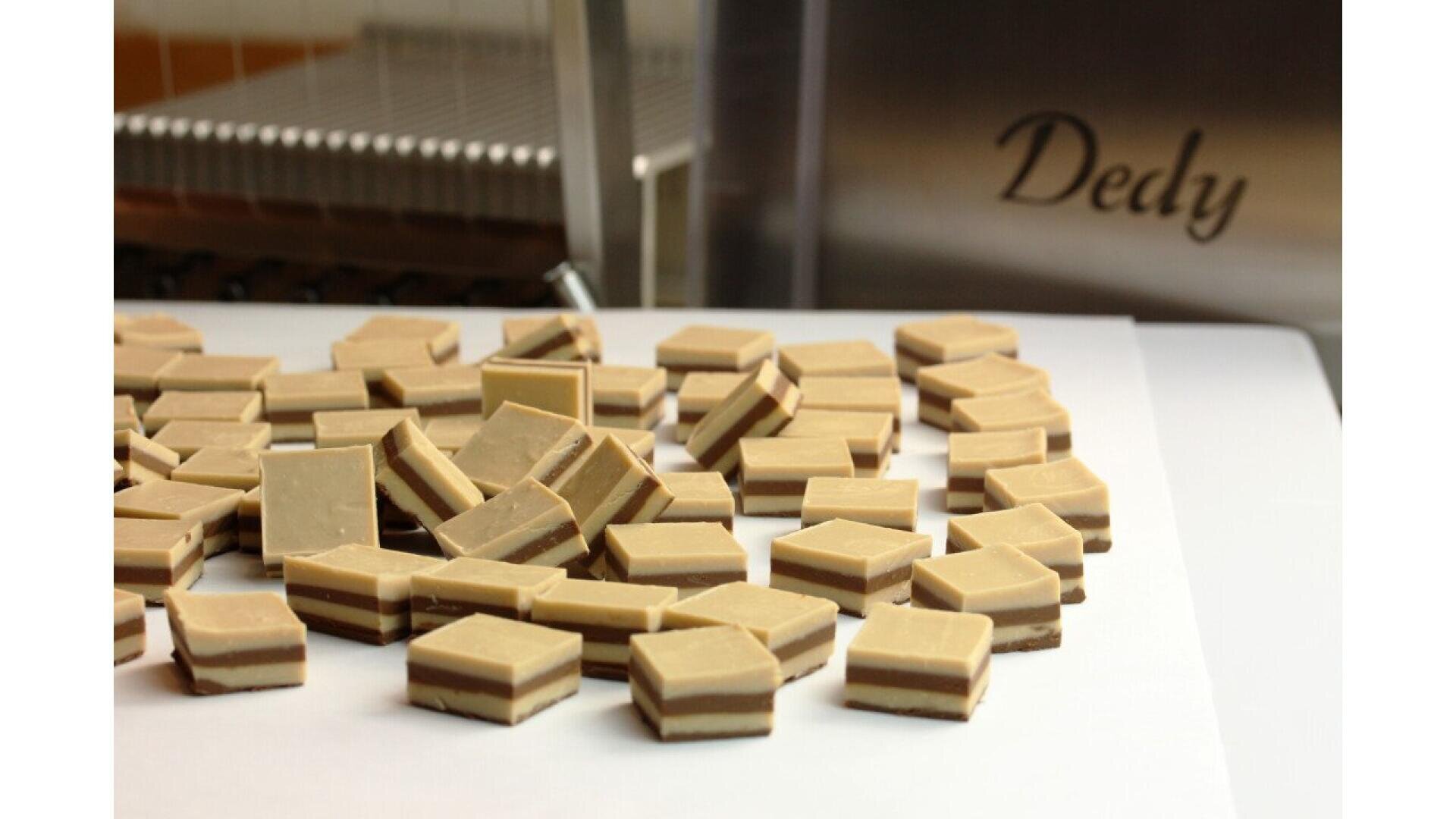A pile of rectangular, layered chocolates with light and dark layers sits on a white surface. In the background, a metal machine is visible with the word “Dedy” etched on it.
