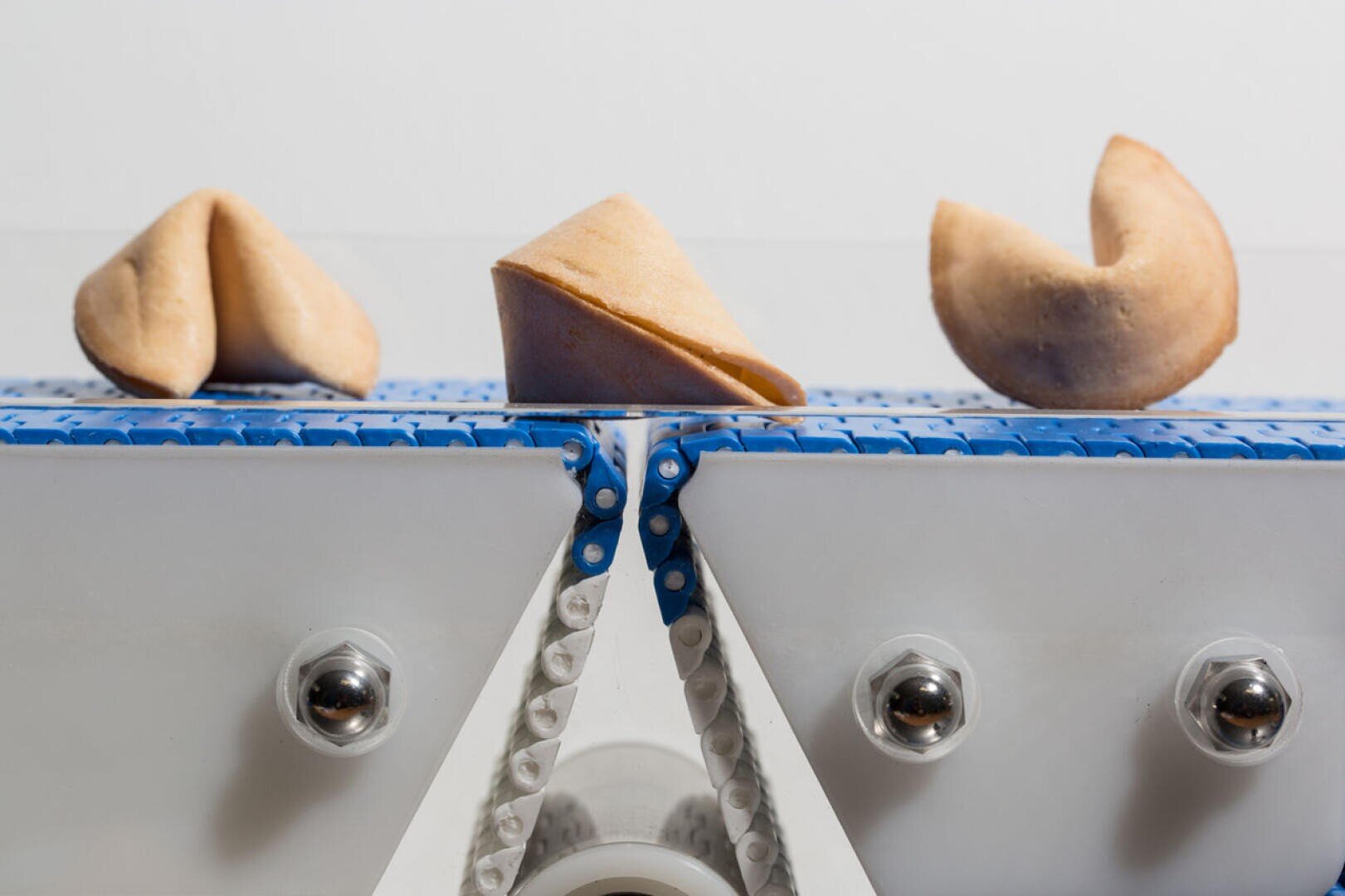 Three fortune cookies are positioned side by side on a blue and white conveyor belt, with a white background. The cookies are in different stages of being folded.