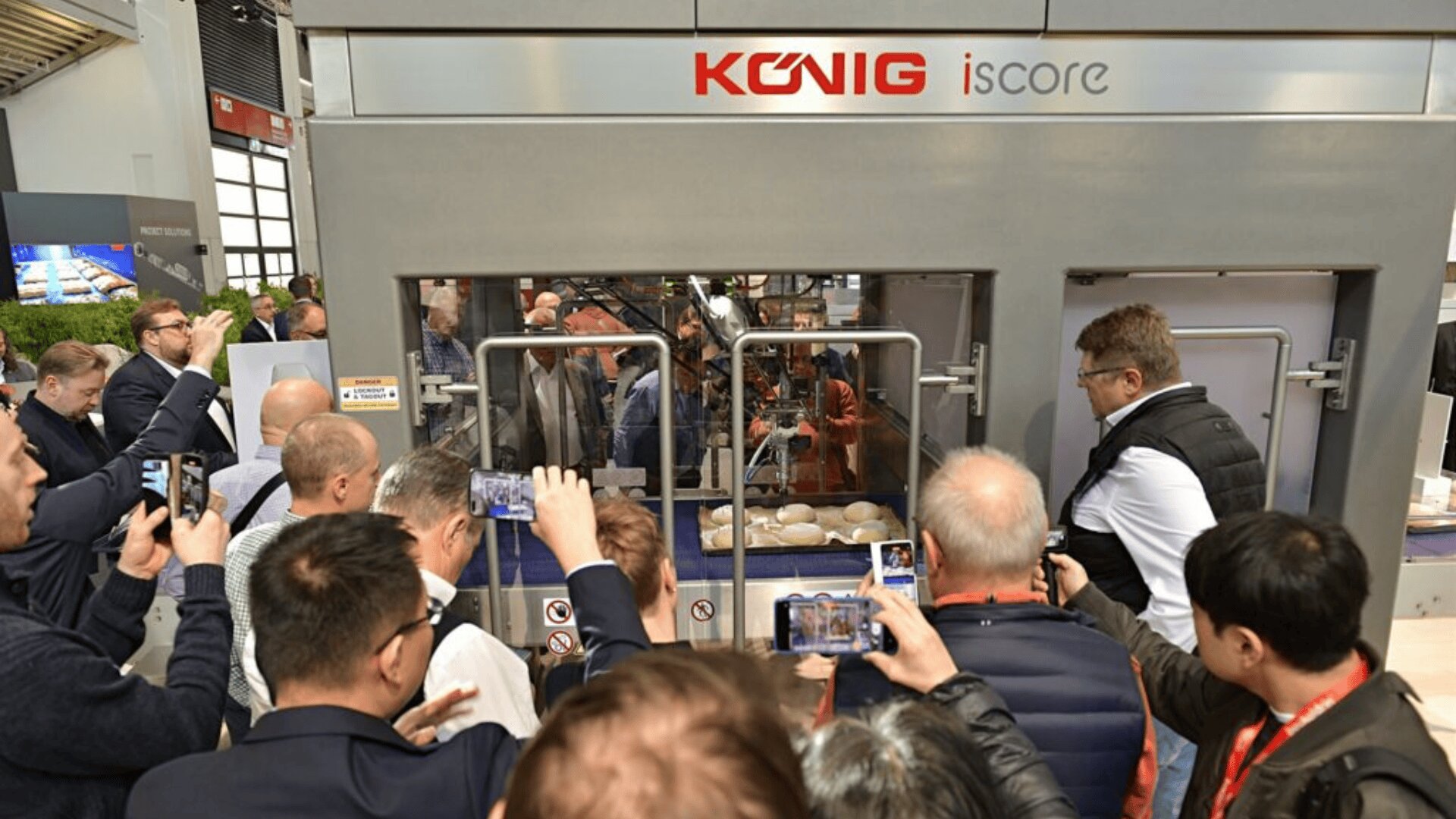 Eine Gruppe von Menschen versammelt sich um eine große Industriemaschine mit der Aufschrift "König iscore" und macht Fotos und Videos, während sie in einem Glasgehäuse auf einer Ausstellung oder Messe Teig oder Brotprodukte verarbeitet.