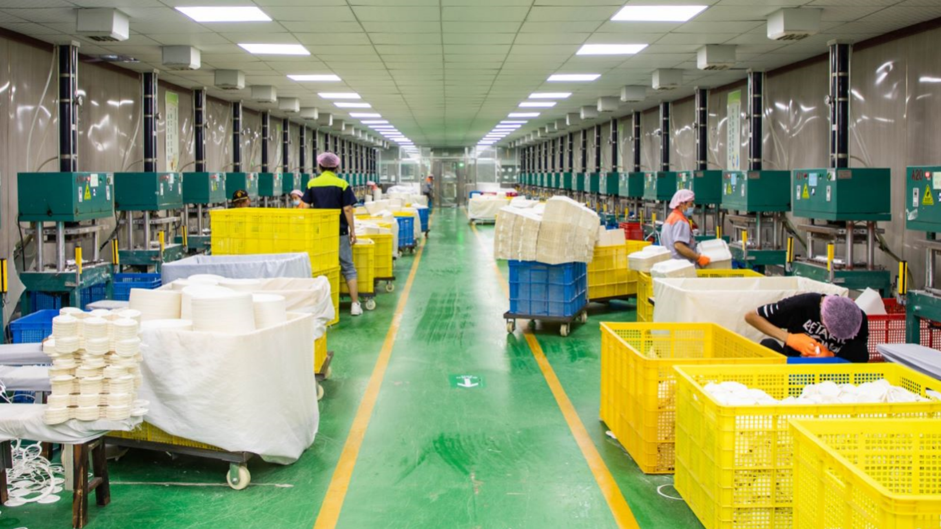 A brightly lit factory interior with workers handling materials at long rows of machines, surrounded by yellow and blue plastic bins and stacks of white products on green flooring.
