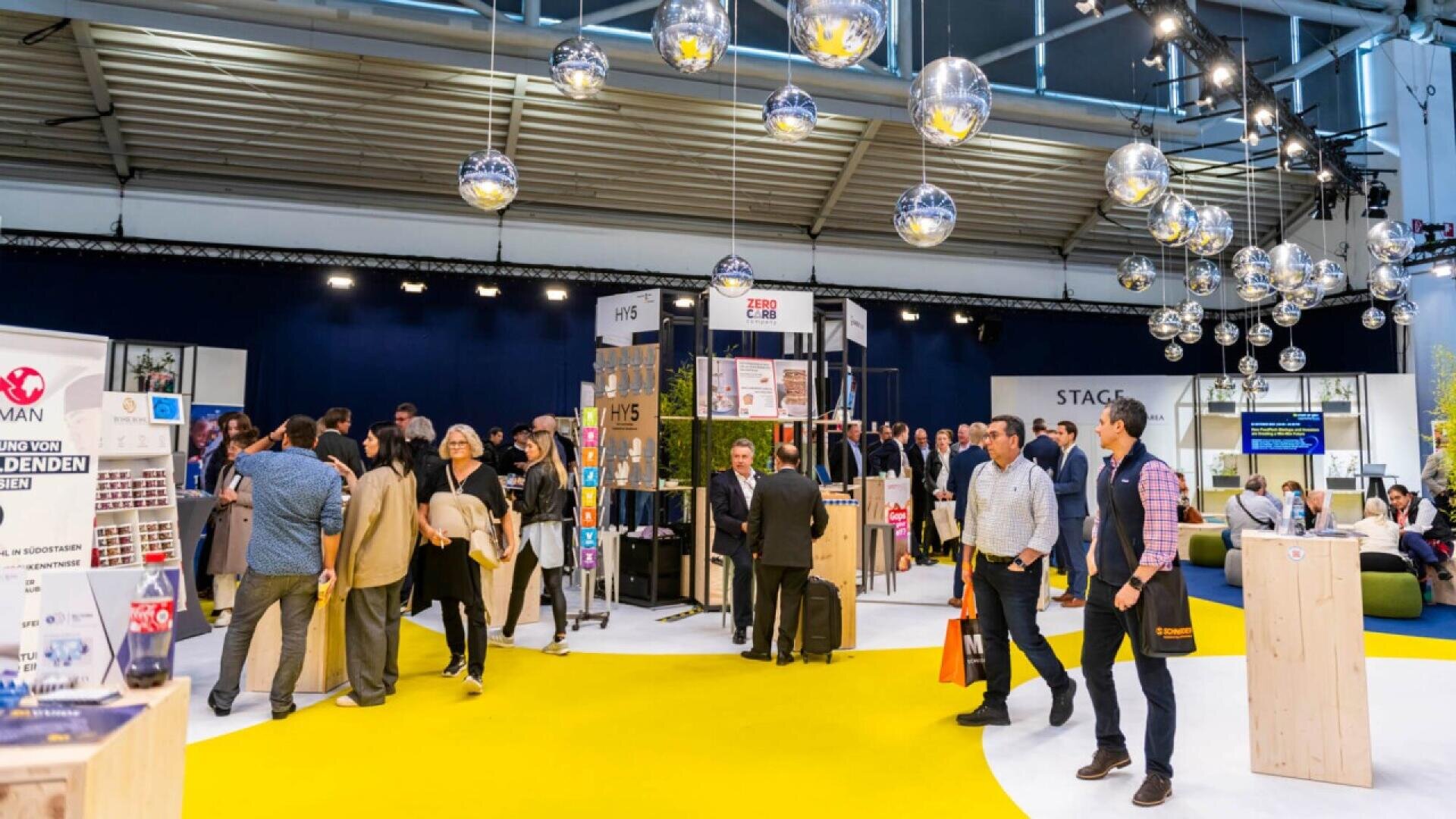 A busy trade show or exhibition hall with people walking and talking near displays and booths under hanging silver globes and bright lighting. The floor is yellow and white, and posters and banners are visible.
