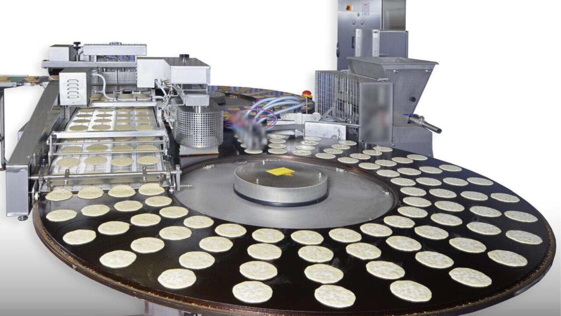A food processing machine produces multiple circular flatbreads or tortillas, placing them evenly on a large round conveyor belt for baking or cooking in an industrial setting.