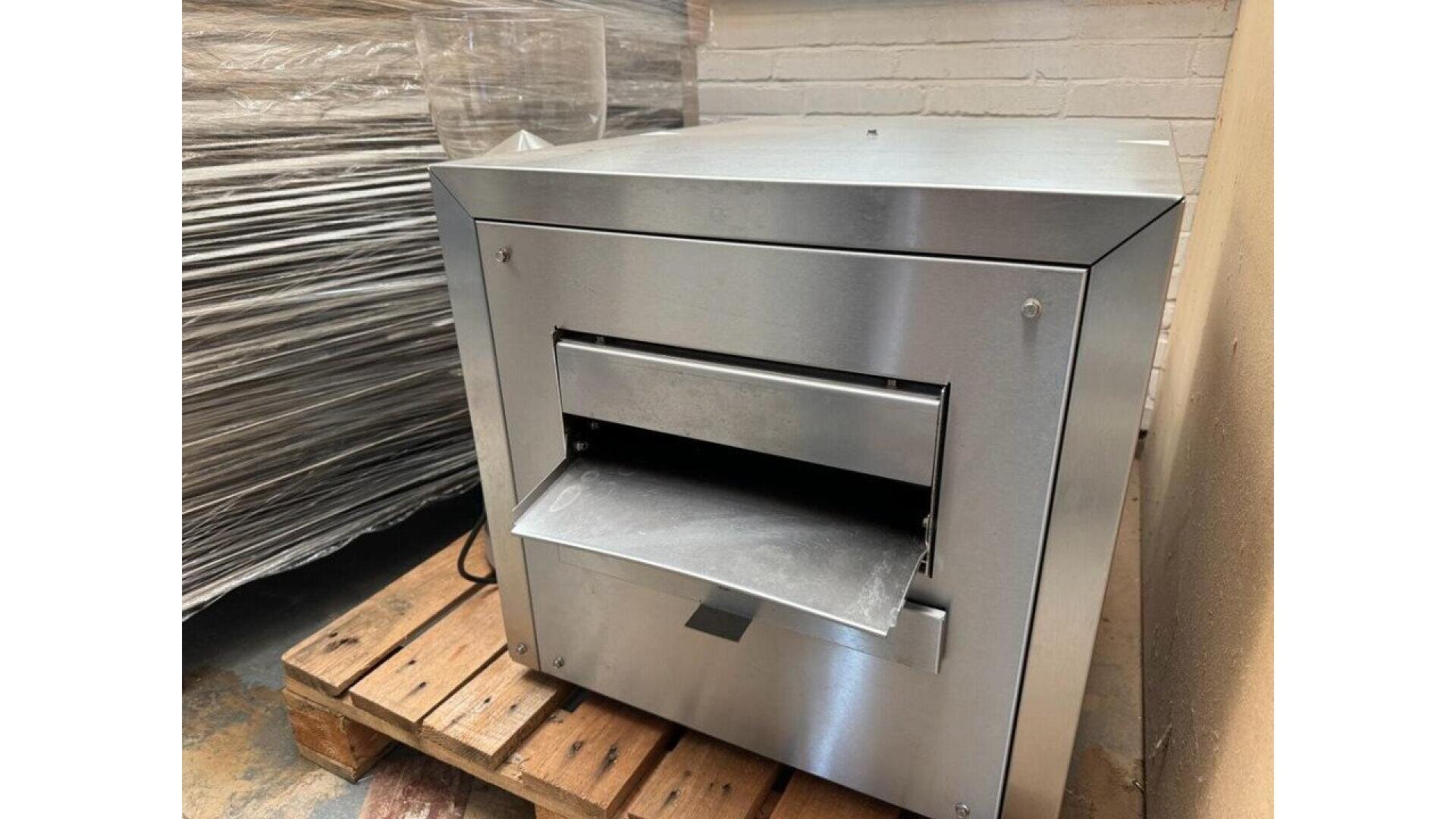 A stainless steel industrial machine sits on a wooden pallet near a brick wall, featuring a rectangular front opening and a slanted metal tray extending outward.