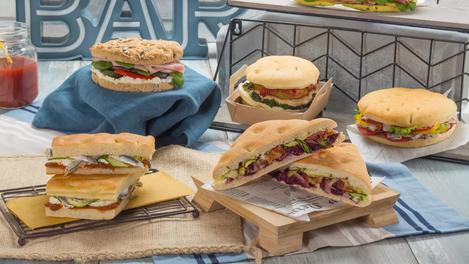 Eine Vielzahl von Gourmet-Sandwiches mit verschiedenen Füllungen wie Gemüse, Grünzeug und Fleisch werden auf Tabletts und Ständern präsentiert, mit dekorativen Requisiten und einem Einmachglas mit Ketchup im Hintergrund.