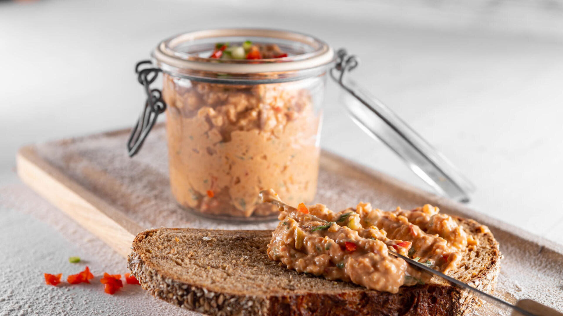 Eine Scheibe Vollkornbrot mit einem cremigen Aufstrich, der gewürfelte rote und grüne Paprika enthält, steht auf einem Holzbrett. Dahinter ein offenes Glas mit mehr von demselben Aufstrich.