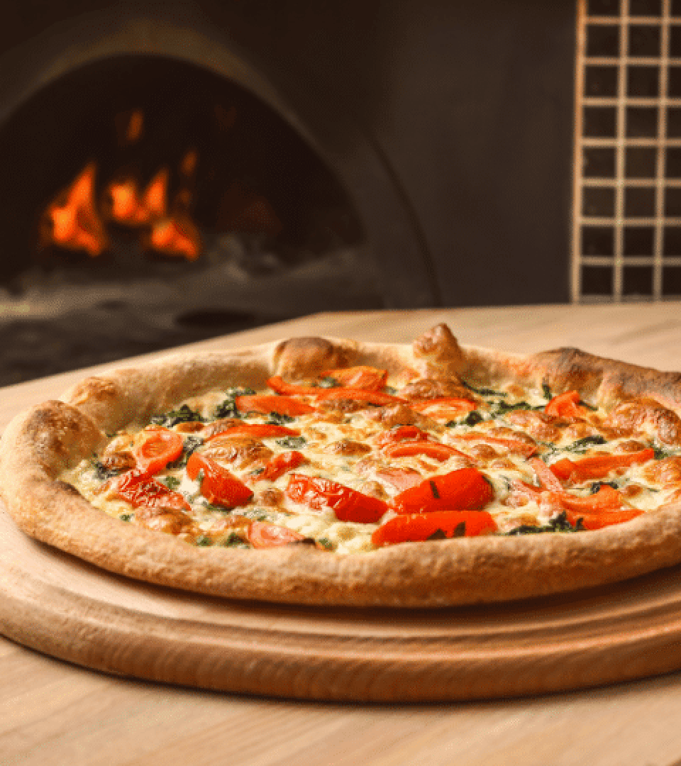 Eine frisch gebackene, mit Tomaten, Käse und Kräutern belegte Pizza liegt auf einem Holzbrett vor einem traditionellen Holzofen, in dem die Flammen sichtbar sind.