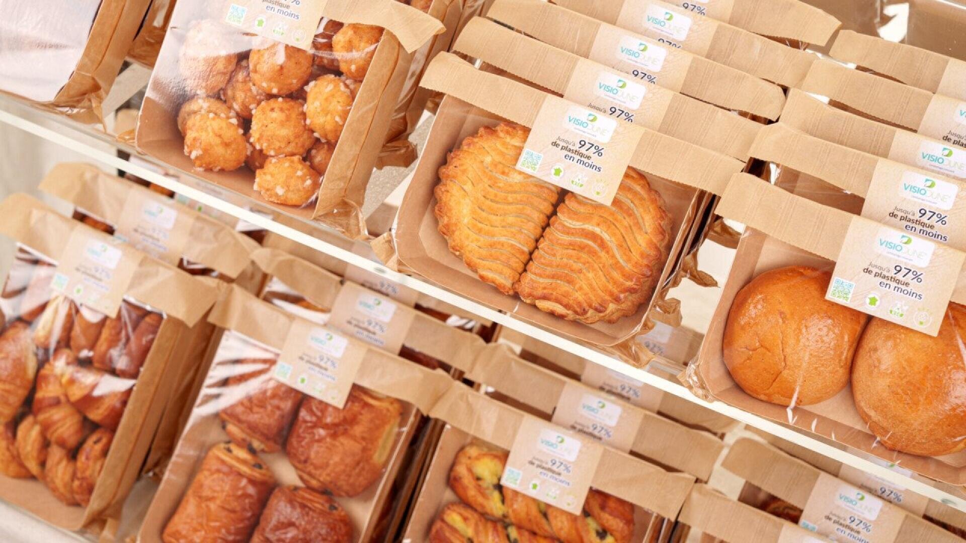 Verschiedene Backwaren, einschließlich Gebäck und Brötchen, werden in Regalen in braunen Papierverpackungen mit durchsichtigen Fenstern und Produktetiketten ordentlich in einer Bäckerei oder einem Geschäft präsentiert.