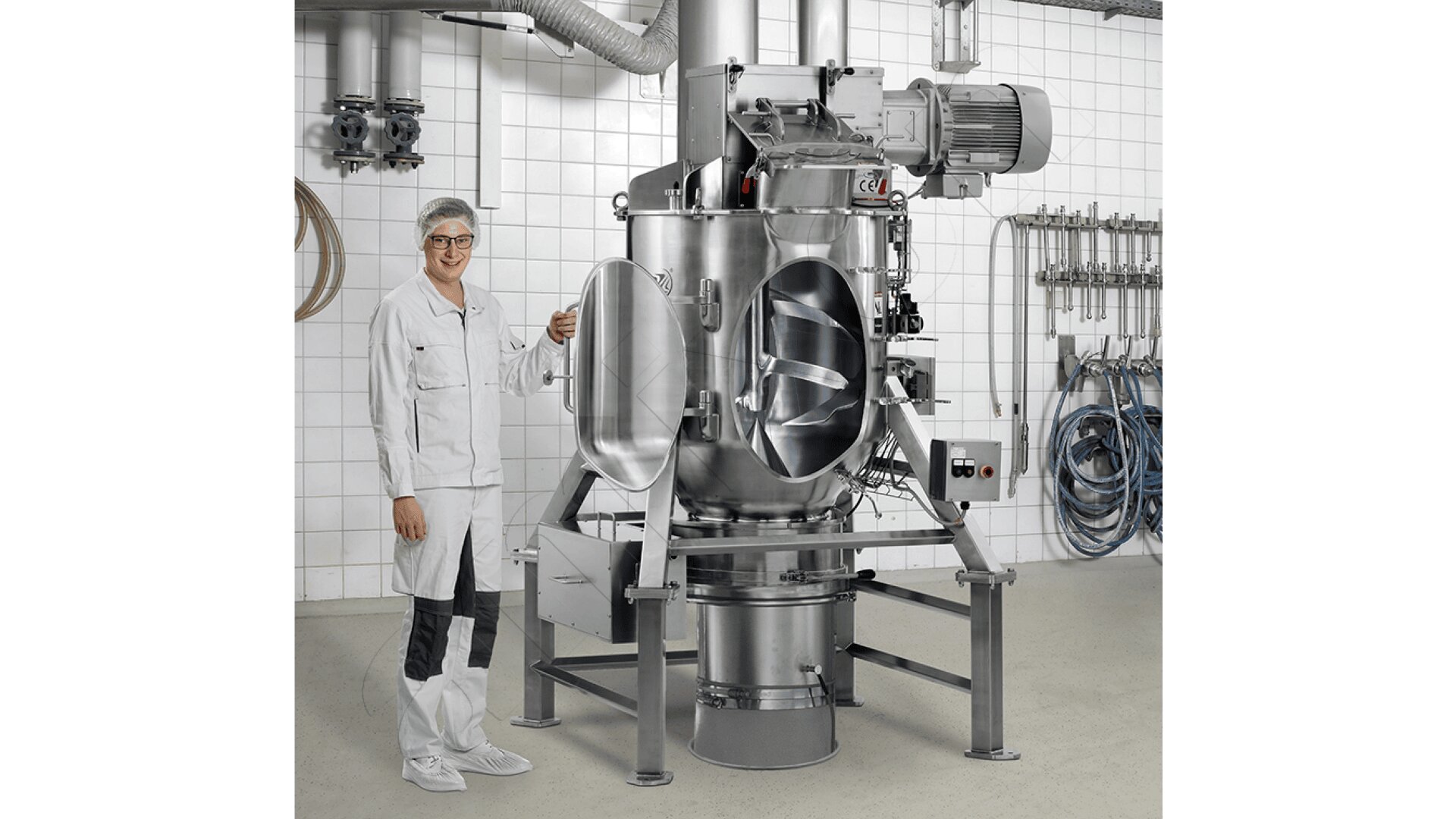 Eine Person in weißer Laborkleidung und einem Haarnetz steht neben einem industriellen Edelstahlmischer in einer sauberen, gefliesten Einrichtung mit verschiedenen Rohren und Schläuchen an der Wand.