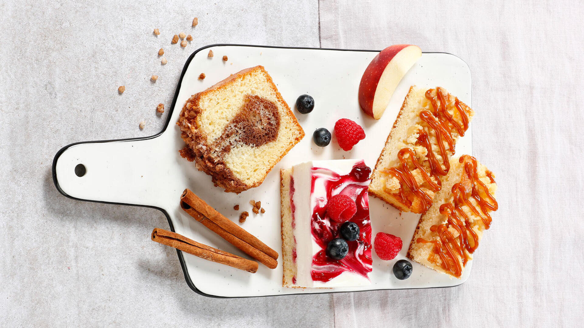 Ein weißes Servierbrett mit Zimtstangen, einer marmorierten Laibkuchenscheibe, Käsekuchen mit Beeren, mit Karamell bestreuten Riegeln, Apfelscheiben, Blaubeeren, Himbeeren und Streuseln auf einer hellen Unterlage.