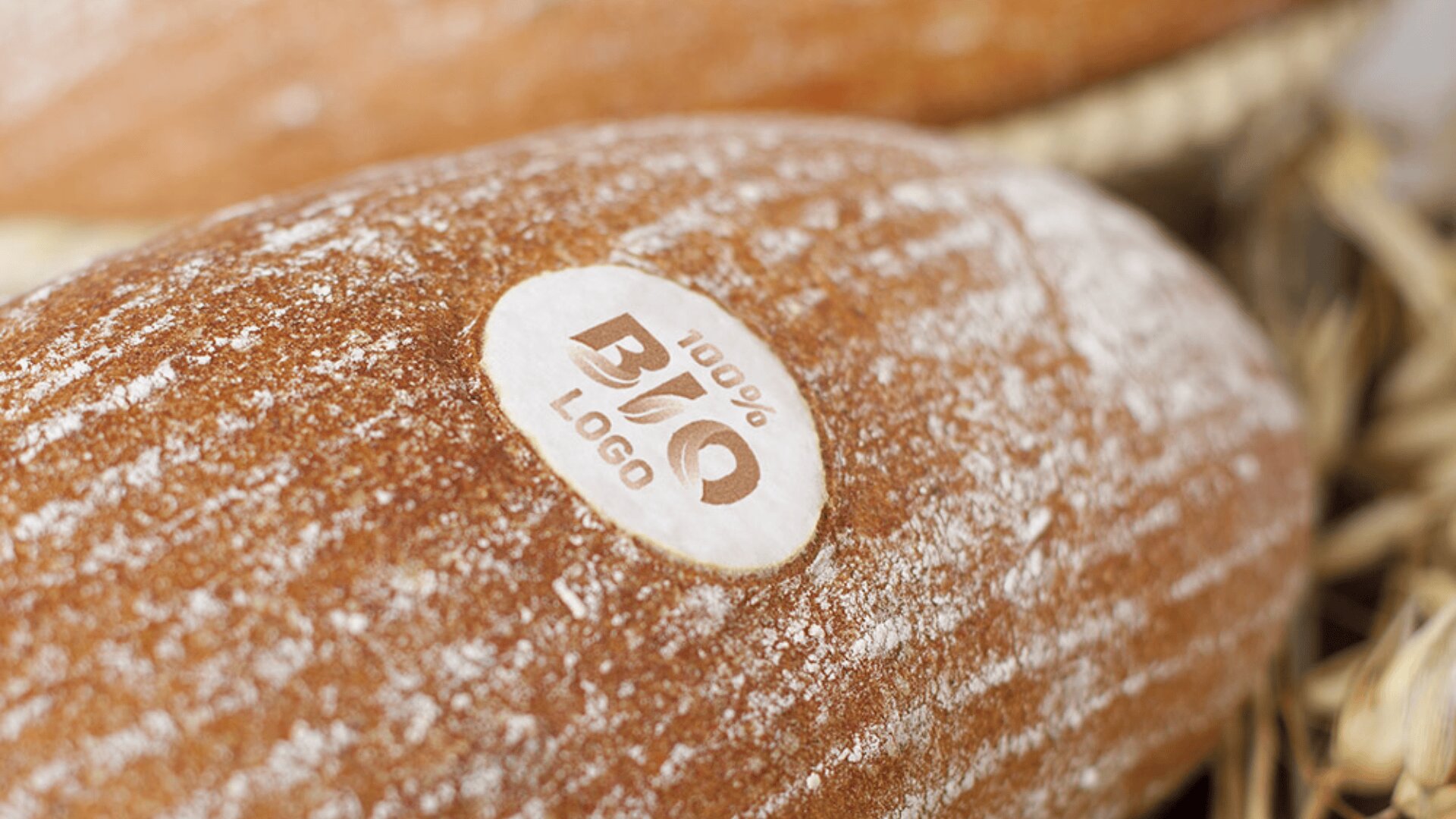 Close-up of a loaf of bread with a round white label that reads 100% BIO LOGO placed on its crust. The bread has a dusting of flour and sits among other loaves with a golden-brown color.